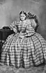 Porträtt av ung kvinna, Anna Carolina Östrand (född 1845) sittande vid bord iklädd rutig klänning, krinolin, med puffärmar och vit krage.