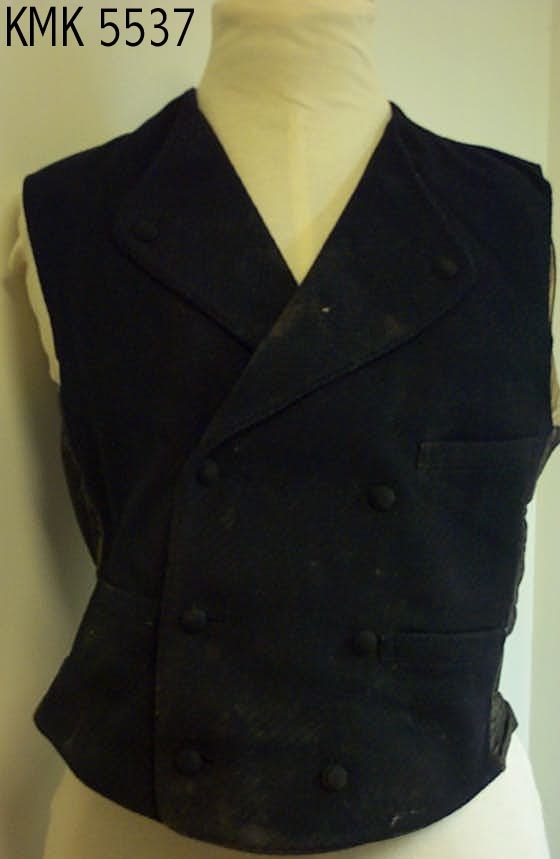 Herrevest med slag, 3 stikklommer og spenne i ryggen. Bakdelen av vesten er i tynnere bomullstoff.  
Vesten lukkes med stofftrukne knapper.