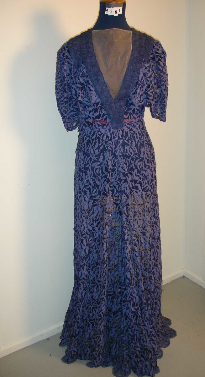 Selskapskjole med korte ermer og kapper på overdelen. Skreddersydd i mørk blå blonderstoff med 11 cm plaststoff sydd på innersiden nederst i kanten (for at skjørtet skal henge penere). Lengde 140 cm. Lukking med trykk-knapper.