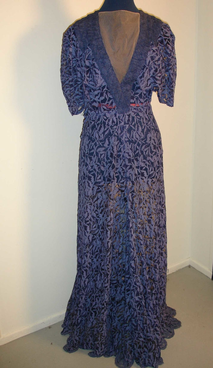 Selskapskjole med korte ermer og kapper på overdelen. Skreddersydd i mørk blå blonderstoff med 11 cm plaststoff sydd på innersiden nederst i kanten (for at skjørtet skal henge penere). Lengde 140 cm. Lukking med trykk-knapper.