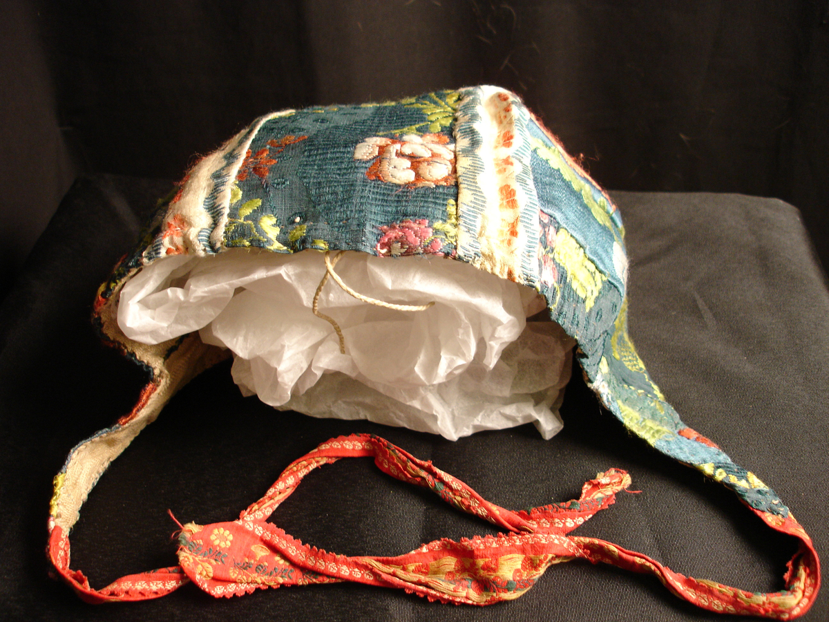 Rynkelue i mønstervevd silke. Lua er satt sammen av mange små biter, og det er flere ulike typer stoff i den. Fôret med linstoff. Den rundede pullen er rynket og sydd til et smalt pannestykke som går over panna og ender i et knytebånd på hver side.