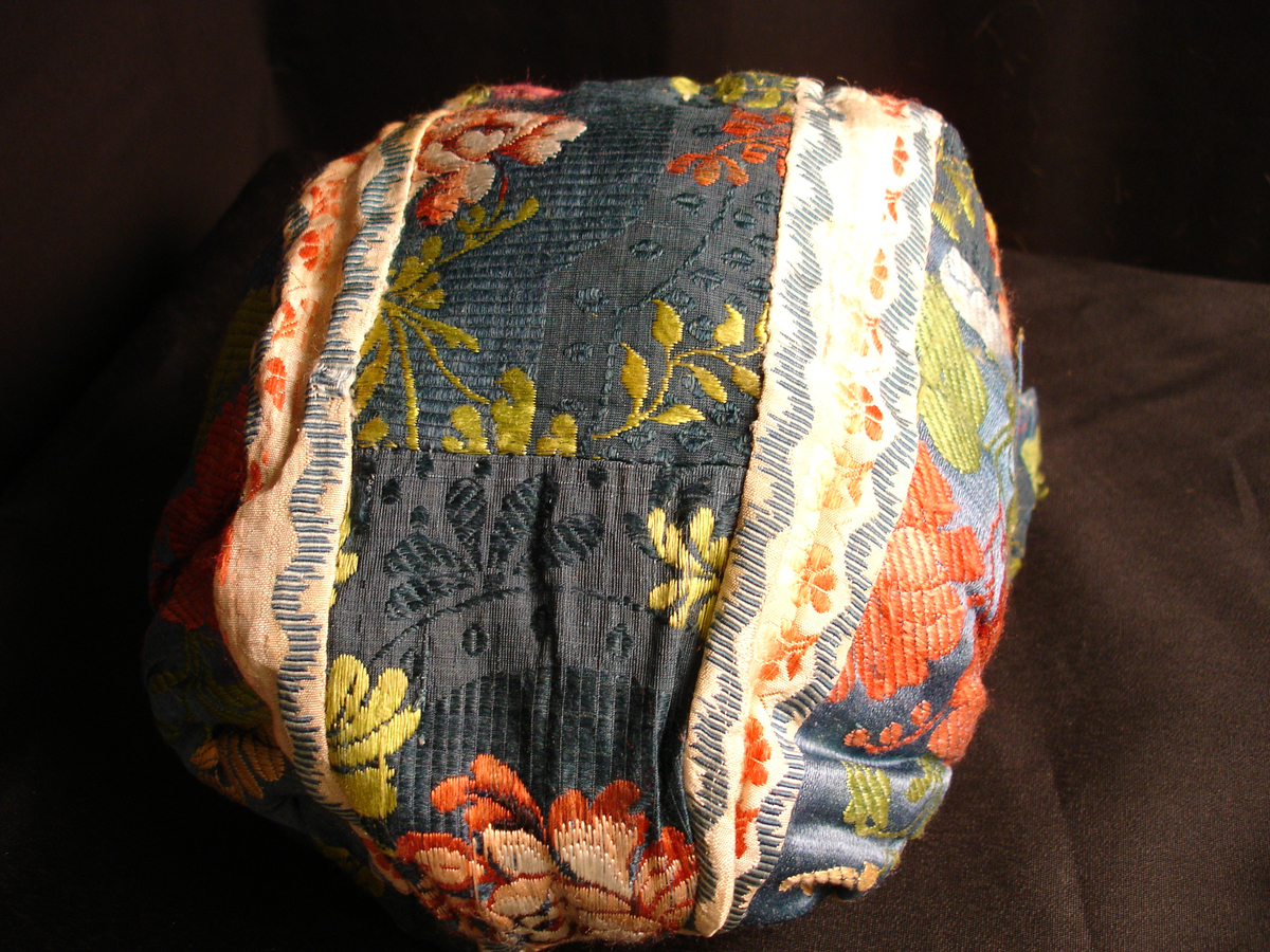 Rynkelue i mønstervevd silke. Lua er satt sammen av mange små biter, og det er flere ulike typer stoff i den. Fôret med linstoff. Den rundede pullen er rynket og sydd til et smalt pannestykke som går over panna og ender i et knytebånd på hver side.