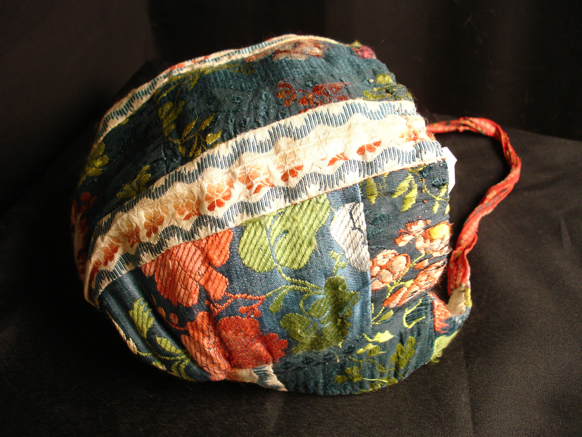 Rynkelue i mønstervevd silke. Lua er satt sammen av mange små biter, og det er flere ulike typer stoff i den. Fôret med linstoff. Den rundede pullen er rynket og sydd til et smalt pannestykke som går over panna og ender i et knytebånd på hver side.