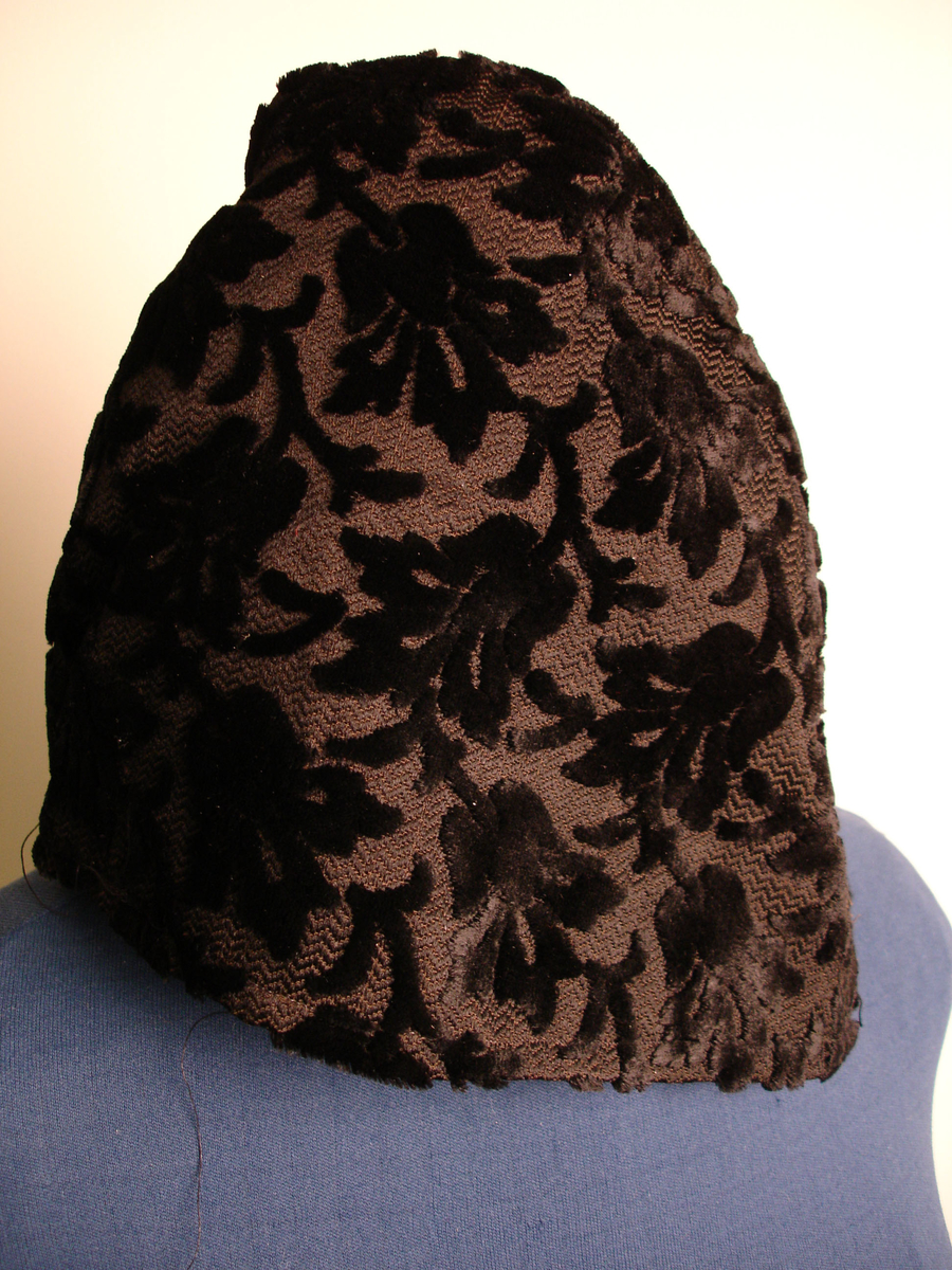 Sort fløyelslue i samme stoff som KMK. 6326. Luen ble antagelig brukt som lue til litt penere bruk. 