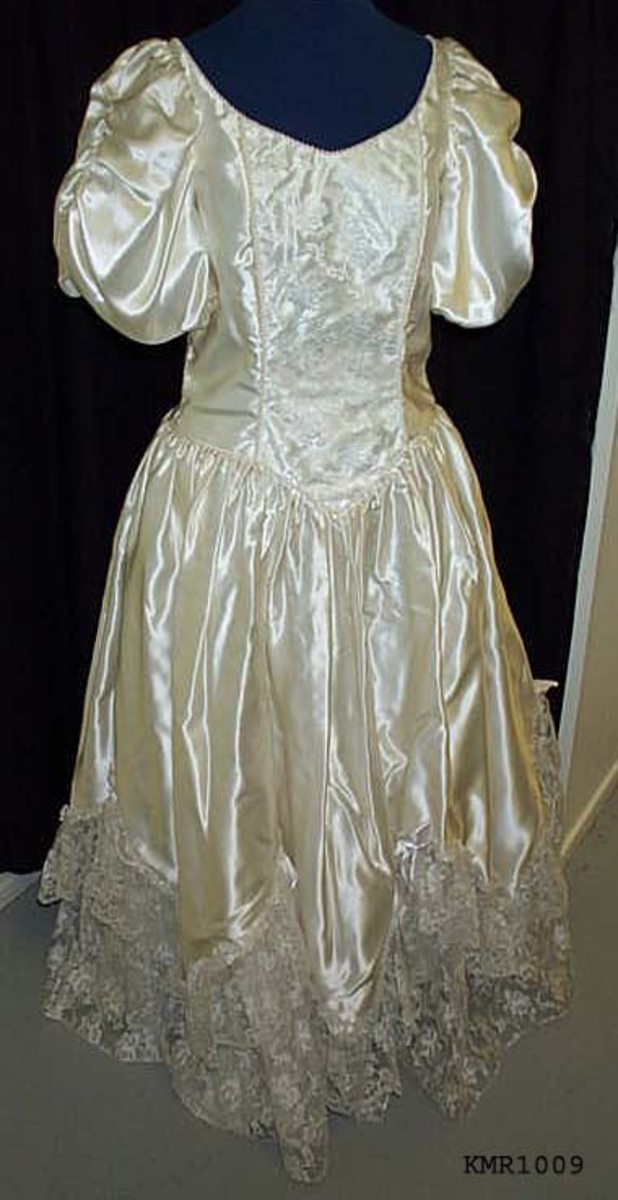  Brudekjole, i hvit silkeaktig stoff, med syntetisk blonderkapper nederst på kjolen 13 cm bred som er sydd i V fasonger, med små sløyfer festet øverst ved hver V, underkjolen er nederste del festet med samme blonderstoffet 50 cm bredde, slik at det ser ut som flere lag kappe. kjolen er avtatt i livet og sømmen er formet som en V i midjen foran, hvor også 15 cm bred blonderstoff er innfelt som pynt i kjolens forstykke, båtformet hals, korte vide ermer 