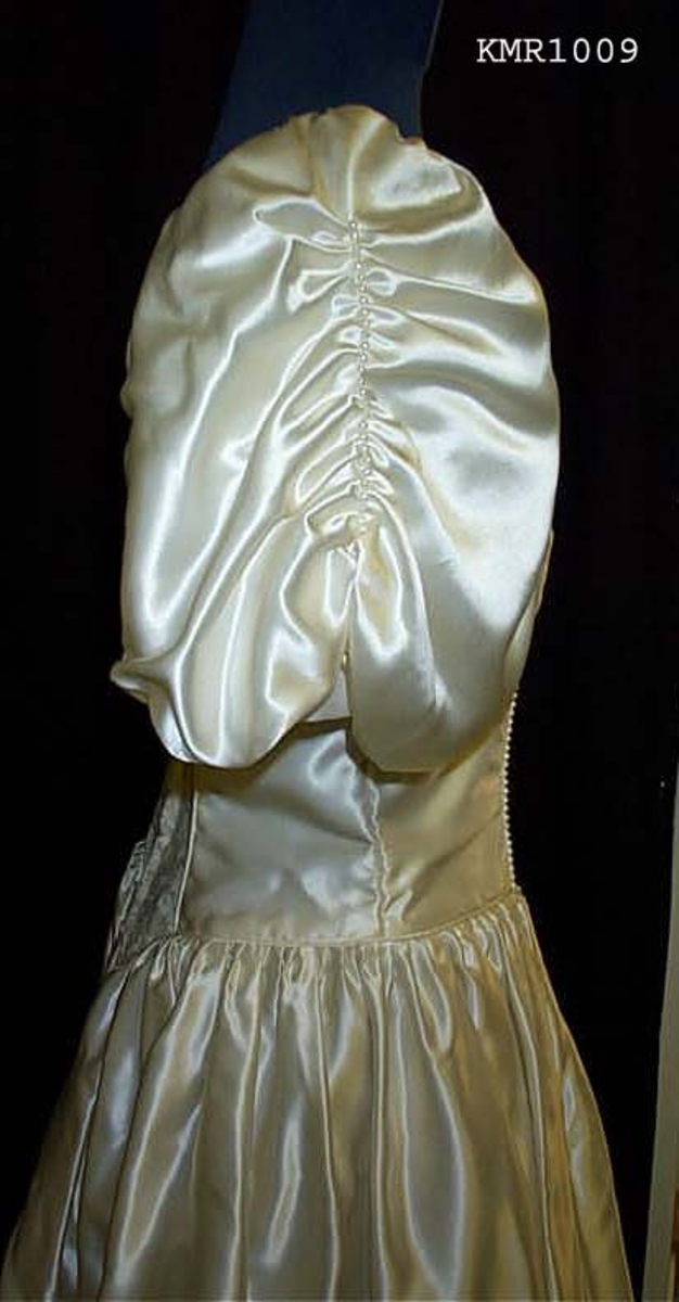  Brudekjole, i hvit silkeaktig stoff, med syntetisk blonderkapper nederst på kjolen 13 cm bred som er sydd i V fasonger, med små sløyfer festet øverst ved hver V, underkjolen er nederste del festet med samme blonderstoffet 50 cm bredde, slik at det ser ut som flere lag kappe. kjolen er avtatt i livet og sømmen er formet som en V i midjen foran, hvor også 15 cm bred blonderstoff er innfelt som pynt i kjolens forstykke, båtformet hals, korte vide ermer 