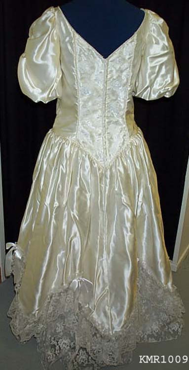  Brudekjole, i hvit silkeaktig stoff, med syntetisk blonderkapper nederst på kjolen 13 cm bred som er sydd i V fasonger, med små sløyfer festet øverst ved hver V, underkjolen er nederste del festet med samme blonderstoffet 50 cm bredde, slik at det ser ut som flere lag kappe. kjolen er avtatt i livet og sømmen er formet som en V i midjen foran, hvor også 15 cm bred blonderstoff er innfelt som pynt i kjolens forstykke, båtformet hals, korte vide ermer 