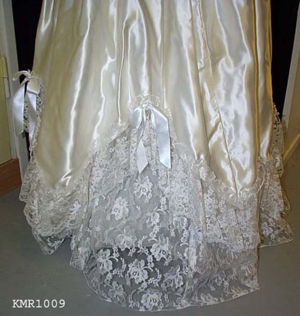  Brudekjole, i hvit silkeaktig stoff, med syntetisk blonderkapper nederst på kjolen 13 cm bred som er sydd i V fasonger, med små sløyfer festet øverst ved hver V, underkjolen er nederste del festet med samme blonderstoffet 50 cm bredde, slik at det ser ut som flere lag kappe. kjolen er avtatt i livet og sømmen er formet som en V i midjen foran, hvor også 15 cm bred blonderstoff er innfelt som pynt i kjolens forstykke, båtformet hals, korte vide ermer 