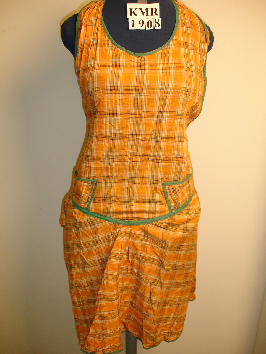 Forkle i rutete flerfarget tekstil og med grønt bånd langs kanter.
Til å tre over hodet, knytes i ryggen. To små pyntelommer i livet.