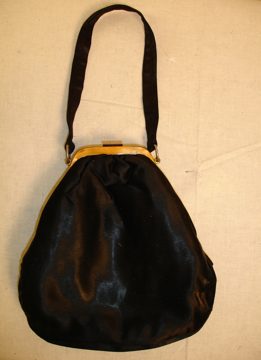 Selskapsveske i svart silkesateng med messinglås (som er noe deffekt) hank, i samme stoff som vesken. Liten lomme på innsiden av vesken.