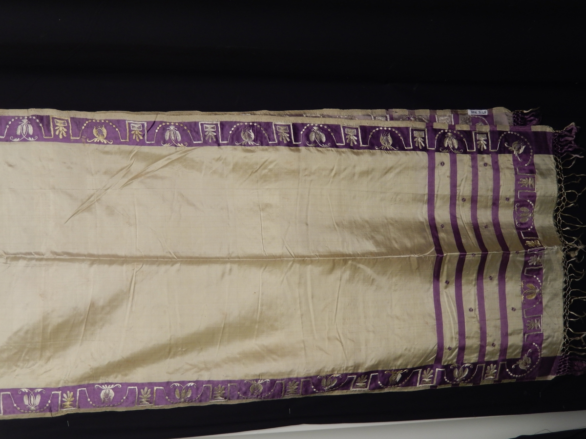 Sjal i silke med frynser. Farge gul med lilla 4 smale striper i endene, bred lilla stripe med broderi i gult på den lilla kanten som er i enden og langs kanten av sjalet. 