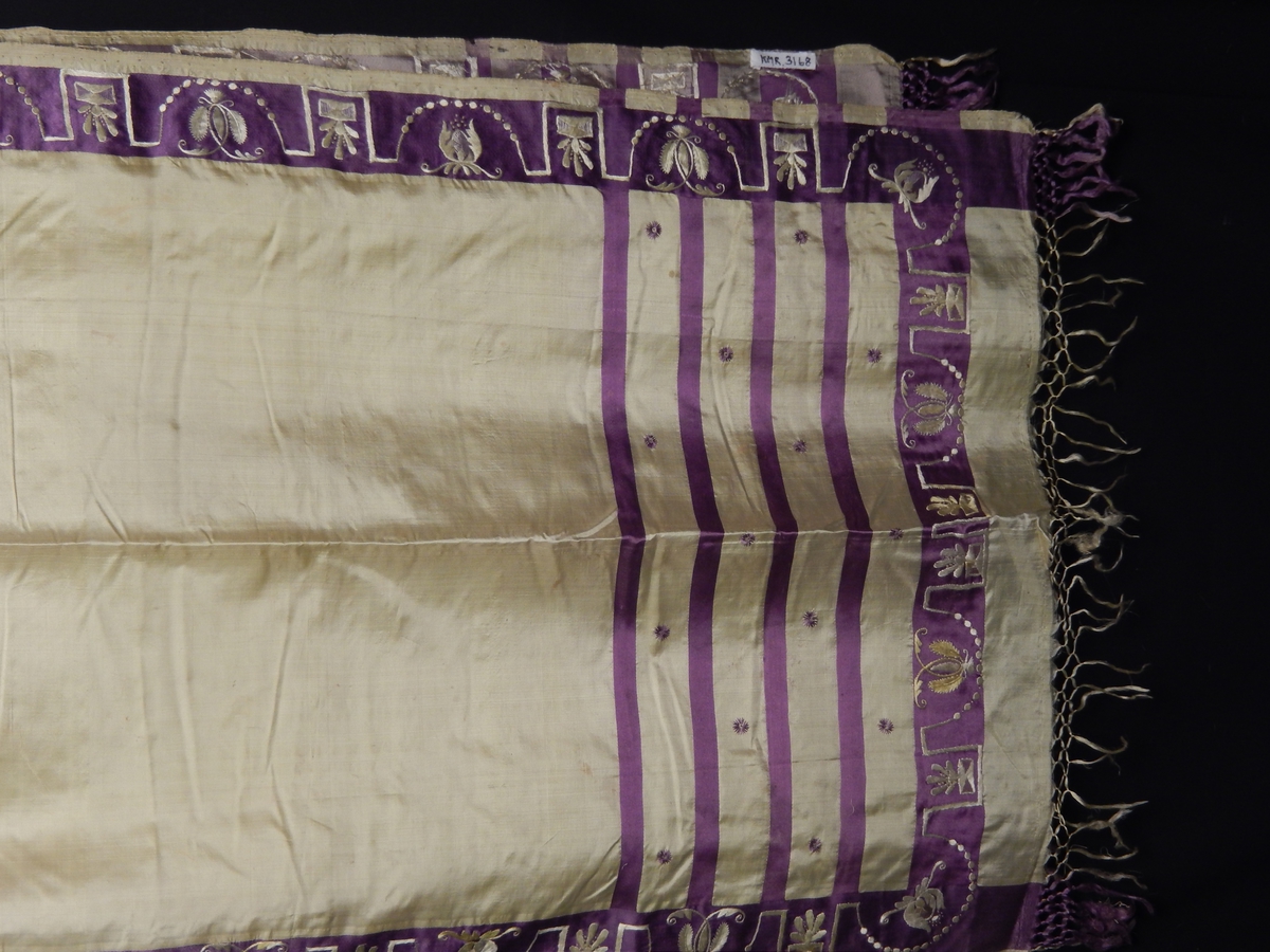 Sjal i silke med frynser. Farge gul med lilla 4 smale striper i endene, bred lilla stripe med broderi i gult på den lilla kanten som er i enden og langs kanten av sjalet. 