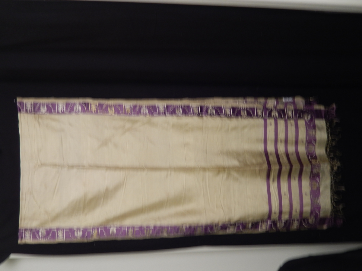 Sjal i silke med frynser. Farge gul med lilla 4 smale striper i endene, bred lilla stripe med broderi i gult på den lilla kanten som er i enden og langs kanten av sjalet. 