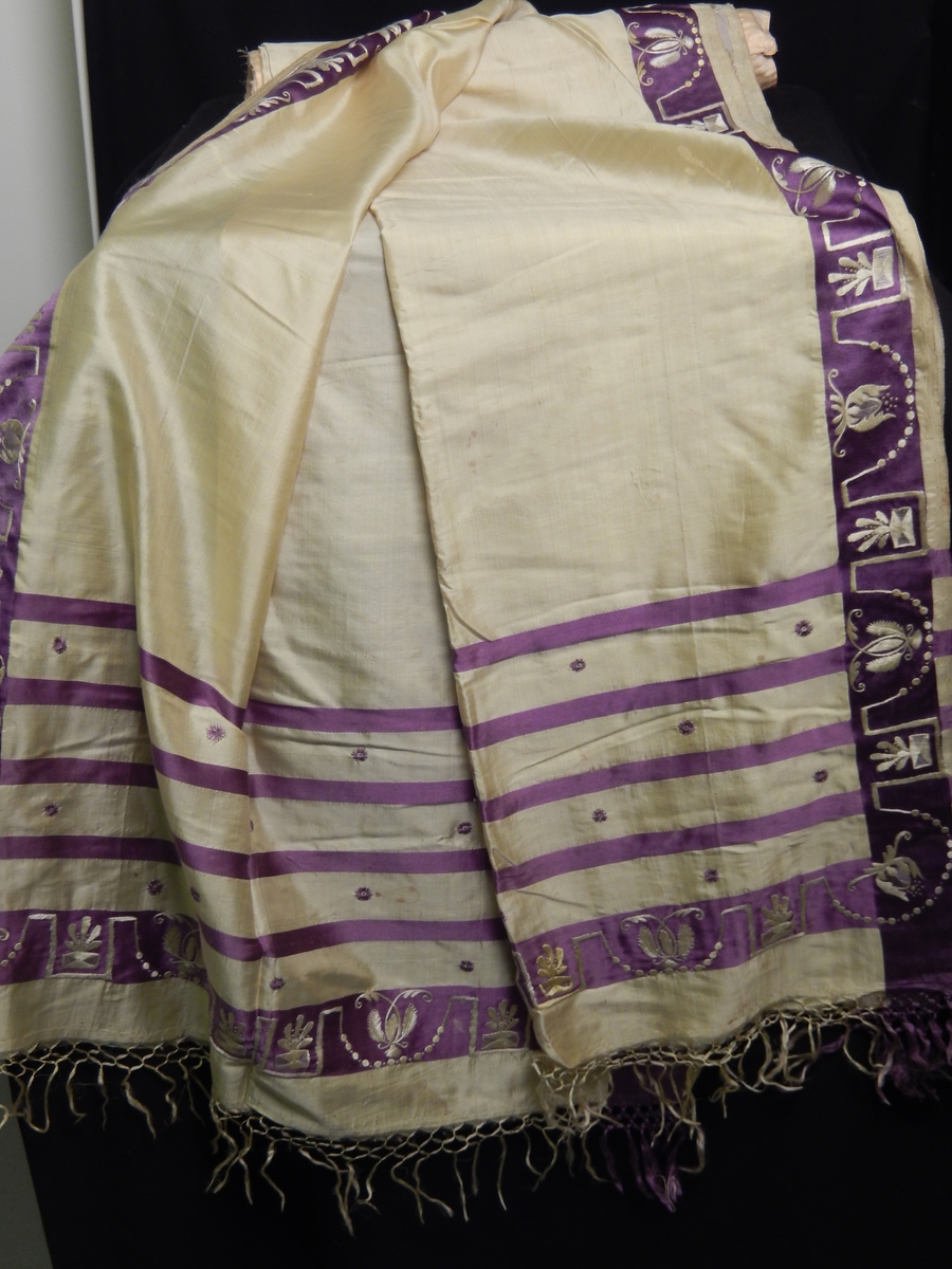 Sjal i silke med frynser. Farge gul med lilla 4 smale striper i endene, bred lilla stripe med broderi i gult på den lilla kanten som er i enden og langs kanten av sjalet. 