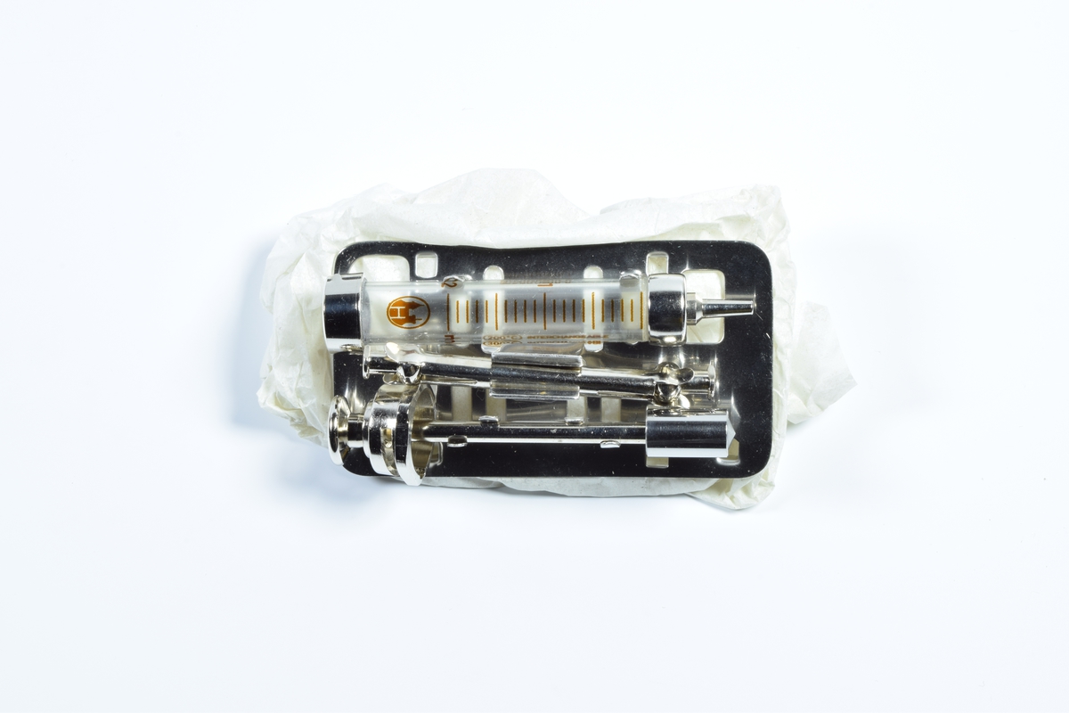 Skrin med lokk i hvit porselen. Skrinet inneholder diverse gjenstander brukt i apotekvirksomhet.

Skrinnet inneholder:

1 grønn eske med "Sterilpakning for kanyler". I esken ligger det 13 små rør i glass og 1 i plast.

1 hvit eske med 8 ampuller med Dolormin til injeksjon.

2 uåpnede pakker med sprøyter, produsert i Tyskland. Eskene er dekorert med et blått kors på en gullfarget bakgrunn og en logo som viser et hus og bostaven H.

2 kvadratiske metallbokser med sprøyter. På lokket til en av boksene er det en logo som viser et hus og bokstaven H.

1 pose med seks munnstykker til nesespylere.

1 lite knivblad med tager og bokstavene A L.

1 pose med flere små stålrør med et lite dråpeformet "håndtak" i en ende.

1 liten pumpe i rød gummi.