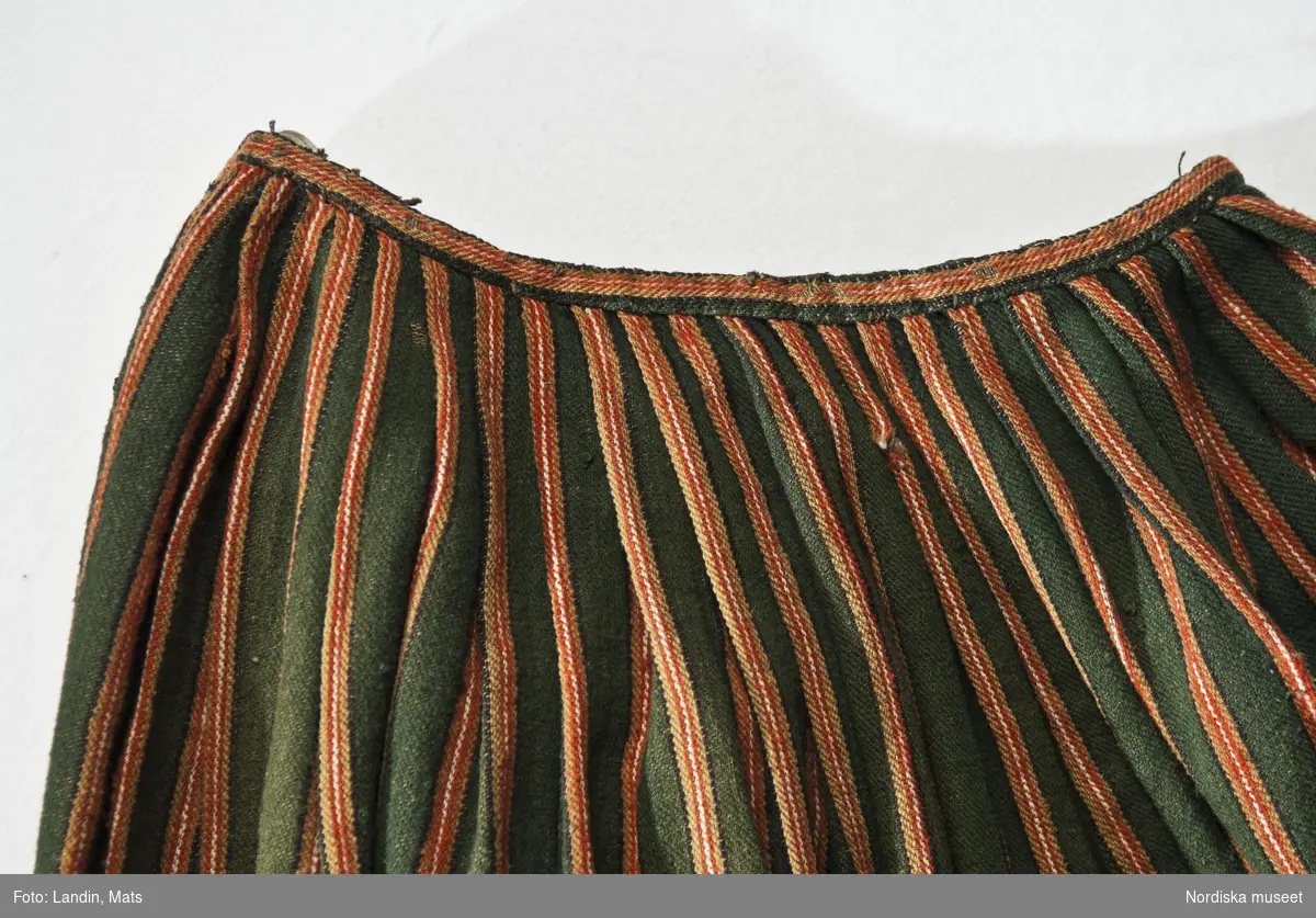 Kjol, helgdagsdräkt, Småland, ca 1800-1825. Halvylle. Kjol av halvylle i kypertbindning, varp av grått lingarn, inslag av entrådigt ullgarn. Veck i midjan lagda så att ränderna ligger överst i vecket. Kjolen saknar veck mitt fram där den är helt slät, sprund i vänster sida under förklädet. Smal fåll utan skoning. Eftersom kvinnorna alltid hade förkläde som dolde framdelen på kjolen, gjordes den oftast slätare och i enklare i mönster och färg just där. Även sprundet sitter oftast under förklädet.