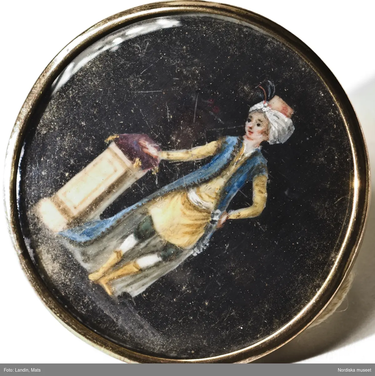 Knappar
Manlig dräkt
Stockholm, eventuellt producerad i Paris, ca. 1780-1790-talet.
Knappar till mansdräkt med miniatyrmålningar på elfenben under glas med förgyllda kanter, gulmetall. 
Motiven föreställer kvinnor och män, alla med tidsenlig dräkt från 1780-1790-talet, mot en svart bakgrund.

Knappar med miniatyrmåleri kom på modet omkring 1775. Handmålade på elfenben, graverade eller ritade. Under 1700-talets sista fjärdedel producerades en hel del sådana knappar i Paris.