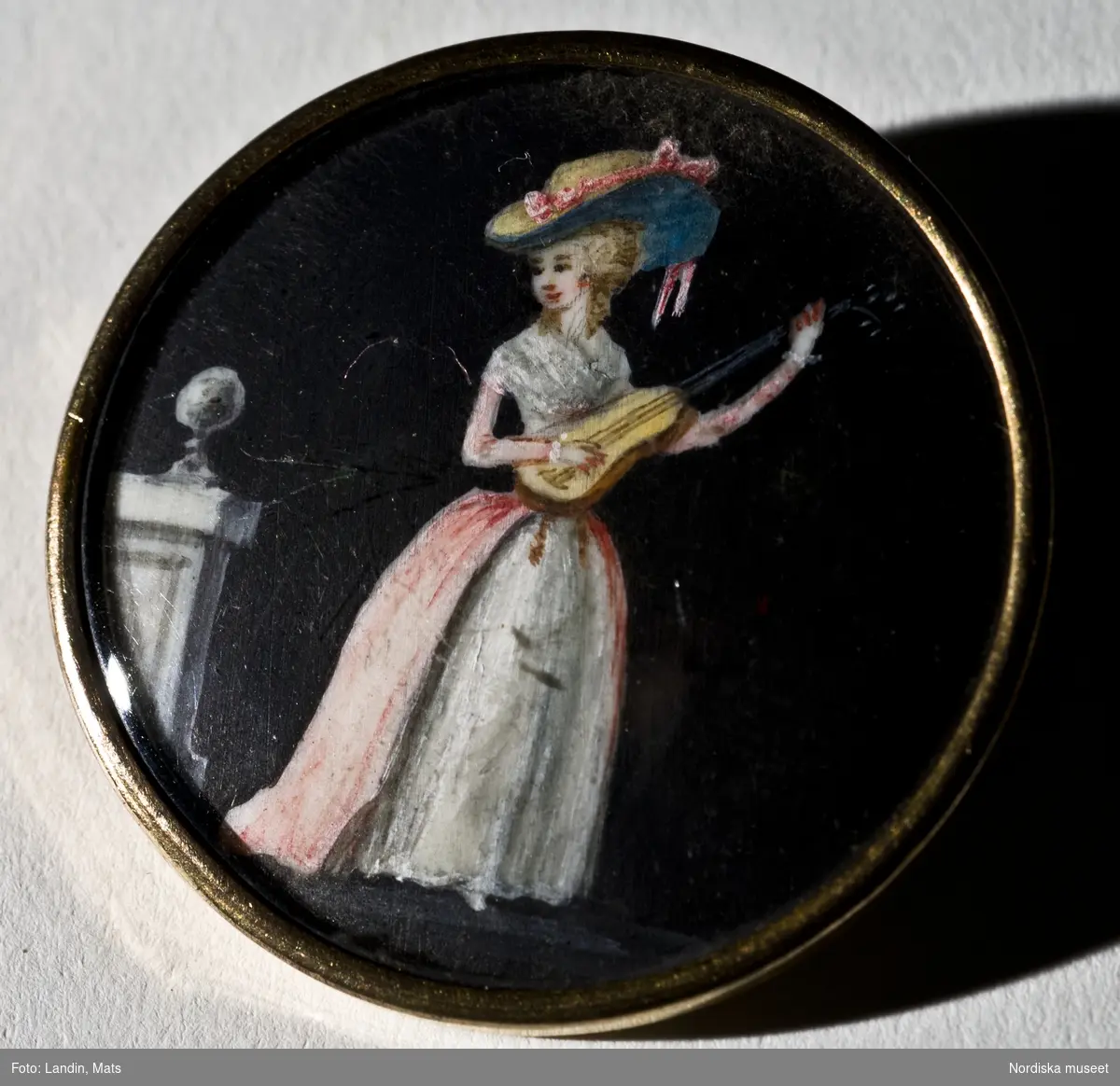 Knappar
Manlig dräkt
Stockholm, eventuellt producerad i Paris, ca. 1780-1790-talet.
Knappar till mansdräkt med miniatyrmålningar på elfenben under glas med förgyllda kanter, gulmetall. 
Motiven föreställer kvinnor och män, alla med tidsenlig dräkt från 1780-1790-talet, mot en svart bakgrund.

Knappar med miniatyrmåleri kom på modet omkring 1775. Handmålade på elfenben, graverade eller ritade. Under 1700-talets sista fjärdedel producerades en hel del sådana knappar i Paris.