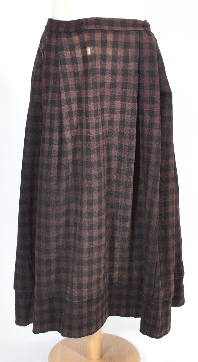 Stakk i samme stoff som liv SM.008023.01; svart og brunrutet ull i hjemmevevd toskaft. Stakken er falmet og slitt med en del hull spredt utover. Sydd sammen for hånd med attersting med svart bomullstråd av fem vevbredder som har jarekanter. Stakken har et 22 cm bredt glatt stykke midt på foran og foldelagt på begge sider. Åpningen er i venstre side og er 21 cm lang. Stakken har fire folder foldelagt mot høyre og seks folder foldelagt mot venstre, 3,5-4,5 cm brede. Midt på bak er det et 13 cm rynket felt med en halv fold over på begge sider. Foldene er tråklet med mørk blå ulltråd, sydd på med attersting med svart bomullstråd og så kastet med faldesting med svart bomullstråd. Linningen er i samme stoff som kjolen og sydd med faldesting med svart bomullstråd deretter med prikkesting med brun lintråd midt på linningen. Lukkes med to metallhekter. Stakken har en 4 cm bred hengefald 11,5 cm fra nedkanten, sydd med faldesting med svart bomullstråd, og en 7,5 cm bred skoning i stripet bomullsstoff sydd med faldesting med svart bomullstråd. Dette er kjøpetøy i fargene beige, rosa, lys blå, blå, mørk blå, hvit og grå. Stakken har en 21 cm lang og 19 cm bred lomme på høyre side i samme stoff som fôret på livet, sydd fast med attersting med ubleket bomullstråd. Stakken har en metallhempe midt på bak på linningen for å hekte på livet og skal brukes utenpå livet.