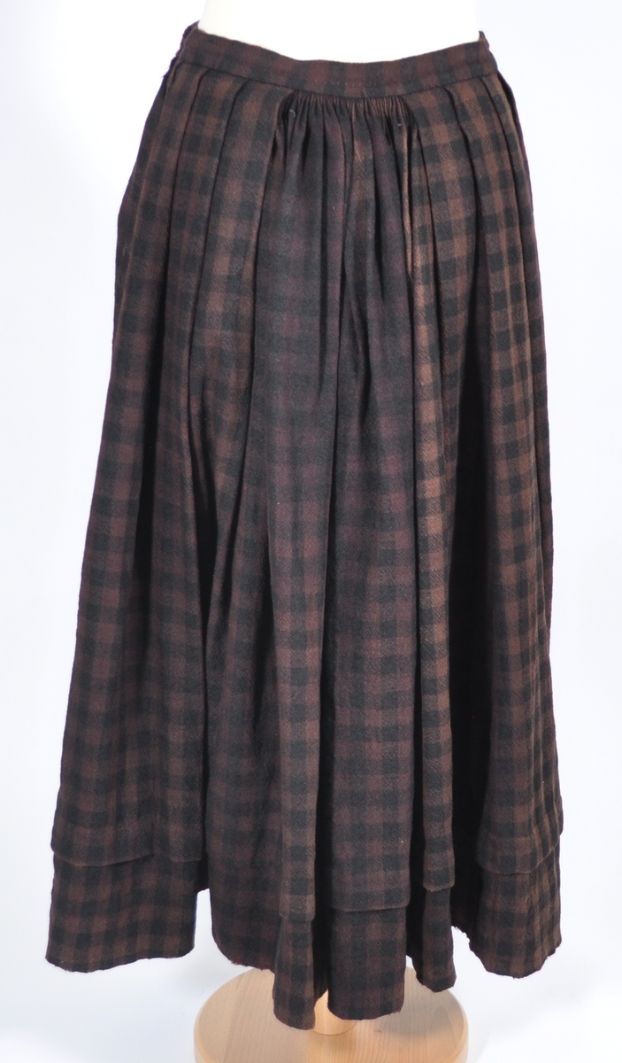 Stakk i samme stoff som liv SM.008023.01; svart og brunrutet ull i hjemmevevd toskaft. Stakken er falmet og slitt med en del hull spredt utover. Sydd sammen for hånd med attersting med svart bomullstråd av fem vevbredder som har jarekanter. Stakken har et 22 cm bredt glatt stykke midt på foran og foldelagt på begge sider. Åpningen er i venstre side og er 21 cm lang. Stakken har fire folder foldelagt mot høyre og seks folder foldelagt mot venstre, 3,5-4,5 cm brede. Midt på bak er det et 13 cm rynket felt med en halv fold over på begge sider. Foldene er tråklet med mørk blå ulltråd, sydd på med attersting med svart bomullstråd og så kastet med faldesting med svart bomullstråd. Linningen er i samme stoff som kjolen og sydd med faldesting med svart bomullstråd deretter med prikkesting med brun lintråd midt på linningen. Lukkes med to metallhekter. Stakken har en 4 cm bred hengefald 11,5 cm fra nedkanten, sydd med faldesting med svart bomullstråd, og en 7,5 cm bred skoning i stripet bomullsstoff sydd med faldesting med svart bomullstråd. Dette er kjøpetøy i fargene beige, rosa, lys blå, blå, mørk blå, hvit og grå. Stakken har en 21 cm lang og 19 cm bred lomme på høyre side i samme stoff som fôret på livet, sydd fast med attersting med ubleket bomullstråd. Stakken har en metallhempe midt på bak på linningen for å hekte på livet og skal brukes utenpå livet.