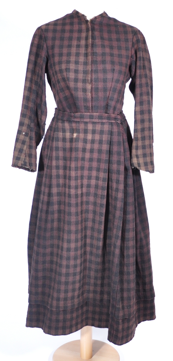 Stakk i samme stoff som liv SM.008023.01; svart og brunrutet ull i hjemmevevd toskaft. Stakken er falmet og slitt med en del hull spredt utover. Sydd sammen for hånd med attersting med svart bomullstråd av fem vevbredder som har jarekanter. Stakken har et 22 cm bredt glatt stykke midt på foran og foldelagt på begge sider. Åpningen er i venstre side og er 21 cm lang. Stakken har fire folder foldelagt mot høyre og seks folder foldelagt mot venstre, 3,5-4,5 cm brede. Midt på bak er det et 13 cm rynket felt med en halv fold over på begge sider. Foldene er tråklet med mørk blå ulltråd, sydd på med attersting med svart bomullstråd og så kastet med faldesting med svart bomullstråd. Linningen er i samme stoff som kjolen og sydd med faldesting med svart bomullstråd deretter med prikkesting med brun lintråd midt på linningen. Lukkes med to metallhekter. Stakken har en 4 cm bred hengefald 11,5 cm fra nedkanten, sydd med faldesting med svart bomullstråd, og en 7,5 cm bred skoning i stripet bomullsstoff sydd med faldesting med svart bomullstråd. Dette er kjøpetøy i fargene beige, rosa, lys blå, blå, mørk blå, hvit og grå. Stakken har en 21 cm lang og 19 cm bred lomme på høyre side i samme stoff som fôret på livet, sydd fast med attersting med ubleket bomullstråd. Stakken har en metallhempe midt på bak på linningen for å hekte på livet og skal brukes utenpå livet.