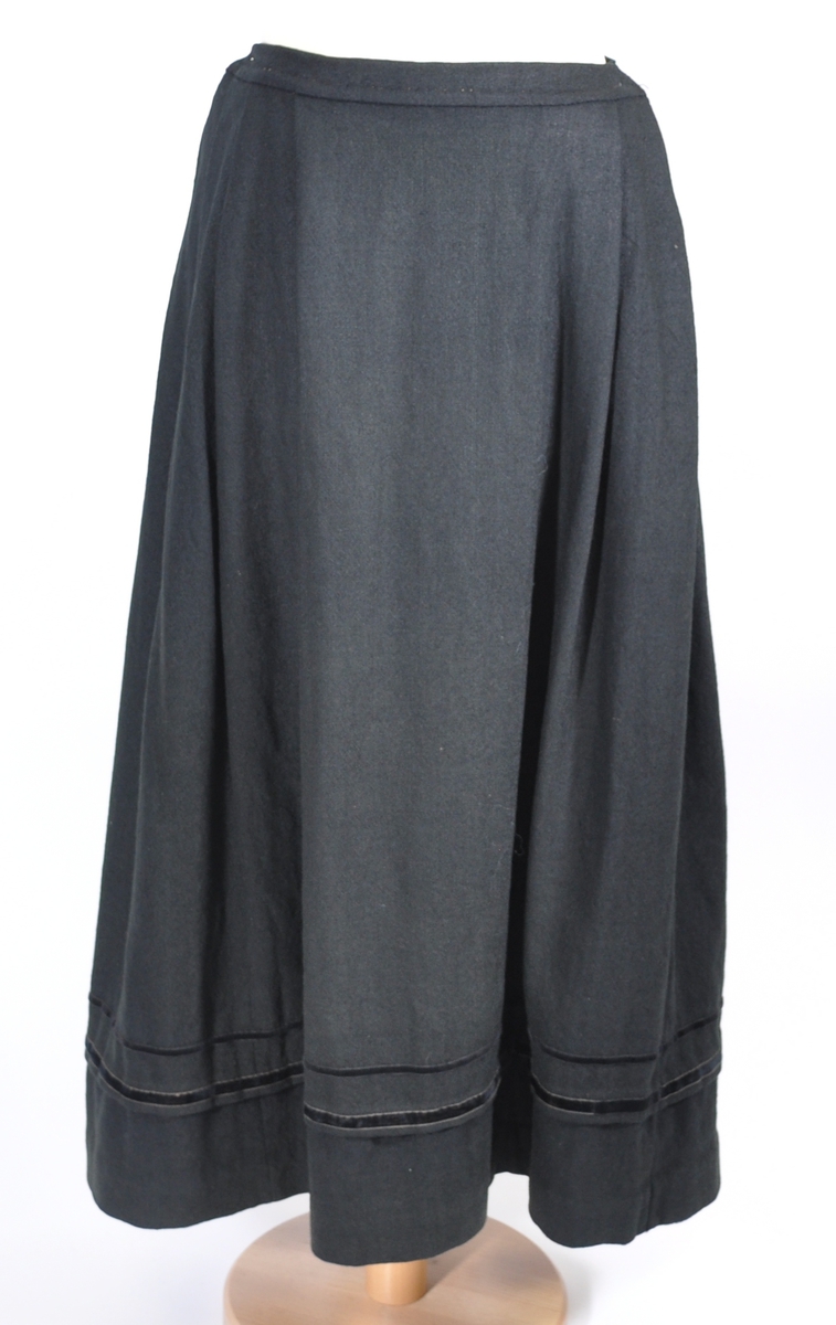 Stakk i samme stoff som liv SM.008024.01; svart ull i hjemmevevd toskaft. Stakken er i god stand med litt slitasje på et pyntebånd. Sydd sammen med maskin med brun bomullstråd av fem vevhøyder med jarekanter. Stakken har et 24 cm bredt glatt stykke midt på foran og foldelagt på begge sider. Åpningen er i venstre side og er 26, 5 cm lang. Stakken har seks folder foldelagt mot høyre og seks folder foldelagt mot venstre, 4-4,5 cm brede. Midt på bak er det et 7,5 cm bredt rynket felt med en halv fold over på begge sider. Foldene er tråklet, sydd på med attersting og så kastet med faldesting med brun bomullstråd. Linningen er i samme stoff som kjolen og 3 cm bred, sydd med faldesting med brun bomullstråd og deretter med prikkesting midt på linningen med samme tråd. Lukkes med to metallhekter. Stakken har en 3 cm bred hengefald 10 cm fra nedkanten, sydd med faldesting med brun bomullstråd. Pyntet nede med to fløyelsbånd som er 0,7 og 1 cm brede, sydd på for hånd i øverste kant av båndet med prikkesting med svart bomullstråd. 0,5 cm bred kostekant i fløyel sydd på med atter- og faldesting for hånd med attersting med svart bomullstråd. Stakken har en 24 cm lang og 17 cm bred lomme på høyre side i samme stoff som fôret på livet, sydd med attersting med svart bomullstråd og festet til stakken med maskin med brun bomullstråd. Stakken har en metallhempe midt på bak på linningen for å henge på livet, og stakken skal brukes ned i livet.