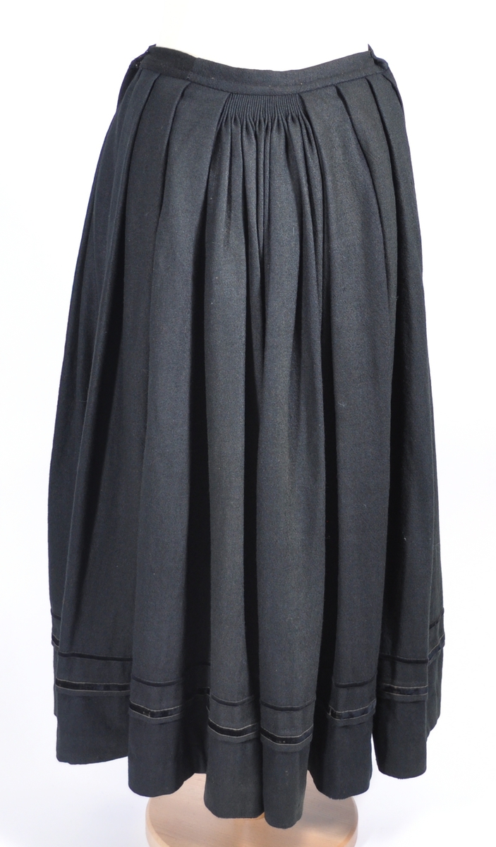 Stakk i samme stoff som liv SM.008024.01; svart ull i hjemmevevd toskaft. Stakken er i god stand med litt slitasje på et pyntebånd. Sydd sammen med maskin med brun bomullstråd av fem vevhøyder med jarekanter. Stakken har et 24 cm bredt glatt stykke midt på foran og foldelagt på begge sider. Åpningen er i venstre side og er 26, 5 cm lang. Stakken har seks folder foldelagt mot høyre og seks folder foldelagt mot venstre, 4-4,5 cm brede. Midt på bak er det et 7,5 cm bredt rynket felt med en halv fold over på begge sider. Foldene er tråklet, sydd på med attersting og så kastet med faldesting med brun bomullstråd. Linningen er i samme stoff som kjolen og 3 cm bred, sydd med faldesting med brun bomullstråd og deretter med prikkesting midt på linningen med samme tråd. Lukkes med to metallhekter. Stakken har en 3 cm bred hengefald 10 cm fra nedkanten, sydd med faldesting med brun bomullstråd. Pyntet nede med to fløyelsbånd som er 0,7 og 1 cm brede, sydd på for hånd i øverste kant av båndet med prikkesting med svart bomullstråd. 0,5 cm bred kostekant i fløyel sydd på med atter- og faldesting for hånd med attersting med svart bomullstråd. Stakken har en 24 cm lang og 17 cm bred lomme på høyre side i samme stoff som fôret på livet, sydd med attersting med svart bomullstråd og festet til stakken med maskin med brun bomullstråd. Stakken har en metallhempe midt på bak på linningen for å henge på livet, og stakken skal brukes ned i livet.