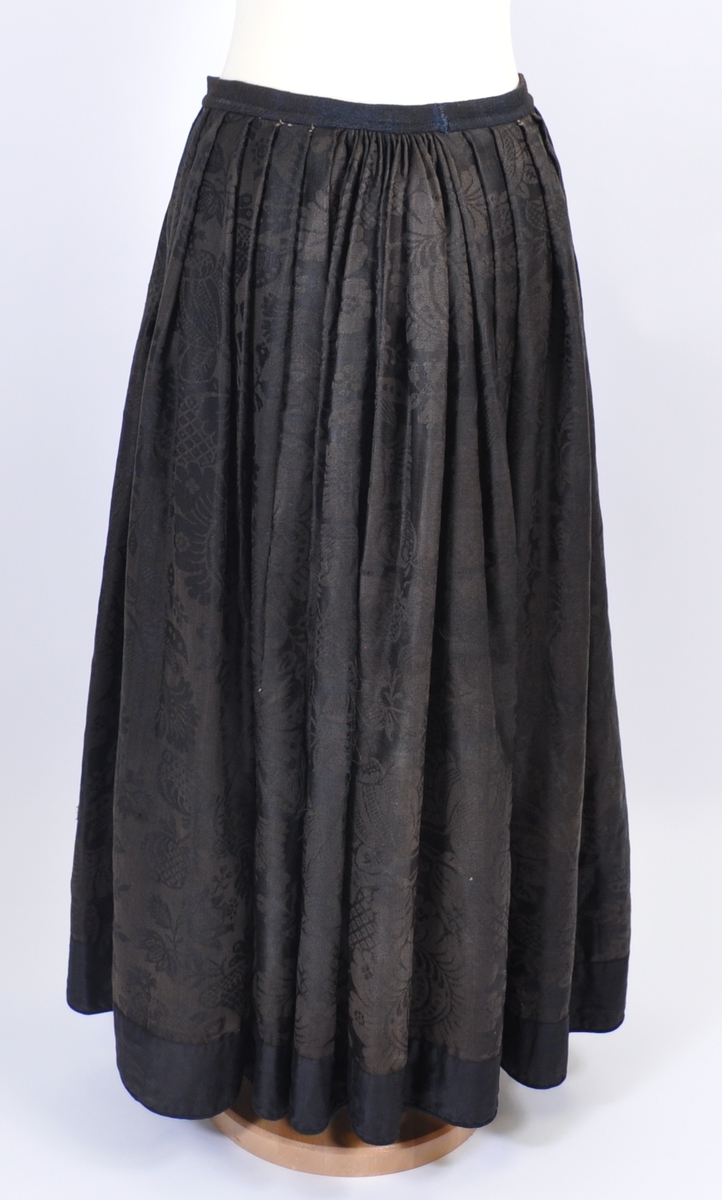 Stakk i brunsvart ulldamask der stoffet i dag er litt falmet, slitt og har noen hull. Stakken er sydd for hånd og maskin, og har blitt bøtt og reparert flere steder. Stakken er skjøtet på og består av sju vevhøyder med jarekant, sydd sammen med attersting med ubleket bomullstråd.  Stakken har et glatt stykke midt på fanghøyden som er 22,5 cm bredt, og foldelagt på begge sider. 28,5 cm lang åpning og en 8 cm bred fold på venstre side av fremstykket. Åpningen har blitt forsterket sekundært med toskafts bomullstøy som er sydd med faldesting med svart sytråd. Stakken har åtte folder foldelagt mot høyre og ti folder foldelagt mot venstre. Midt på bak har stakken et 8 cm bredt felt med rynking. Linningen er av vadmel og trolig sekundær, sydd på med maskin med svart sytråd. Stakken lukkes med to metallhekter og har en metallkrok midt på bak på linningen for å hekte på livet. Stakken har blitt forlenget sekundært med en 8 cm bred kant i svart, glatt bomull, påsydd med maskin med svart sytråd. Dette bomullsstoffet ser ut til å være samme stoff som kjoleliv 6.3.15. Stakken har en 7,2 cm høy skoning i trolig hjemmevevd svart og beige fiskebensmønstret bomull sydd på med maskin og faldesting med svart sytråd. Kantet nede med et 0,5 cm bredt svart kjøpe-ullbånd, sydd på med maskin og faldesting med svart sytråd. Stakken har en sekundær lomme sydd inn i mellom faldene på høyre side. Lommen er 16 cm bred og 21 cm lang, sydd med maskin med svart sytråd, og er i samme stoff som skoningen. Stakken har en metallkrok midt på bak på linningen for å henge på livet, og stakken skal brukes utenpå livet.