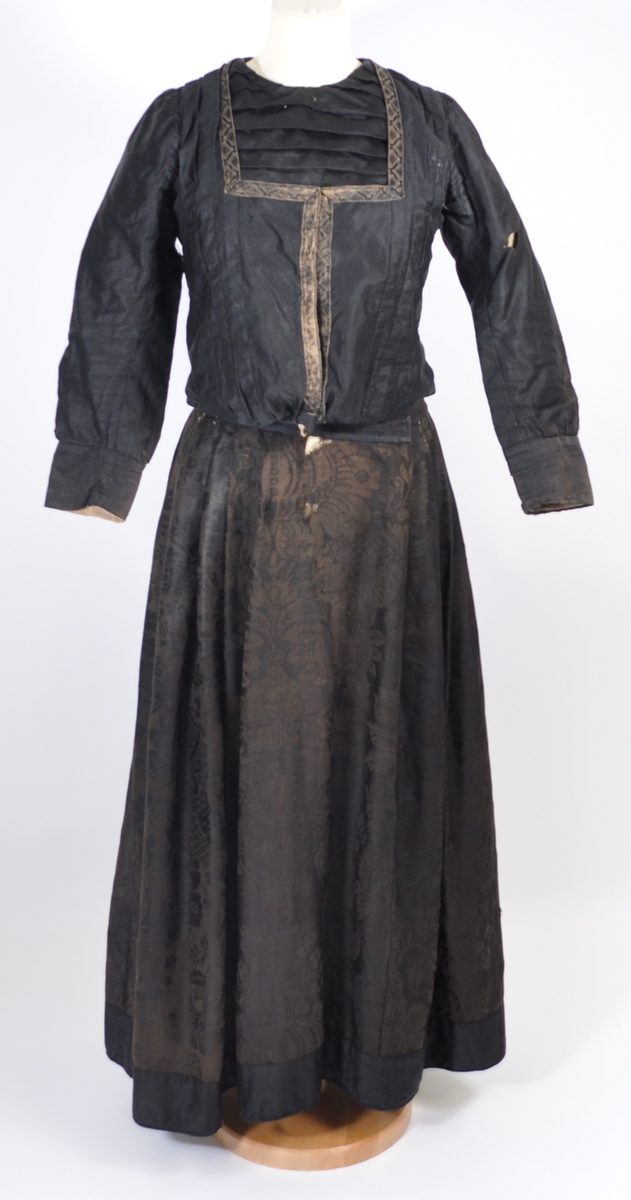 Stakk i brunsvart ulldamask der stoffet i dag er litt falmet, slitt og har noen hull. Stakken er sydd for hånd og maskin, og har blitt bøtt og reparert flere steder. Stakken er skjøtet på og består av sju vevhøyder med jarekant, sydd sammen med attersting med ubleket bomullstråd.  Stakken har et glatt stykke midt på fanghøyden som er 22,5 cm bredt, og foldelagt på begge sider. 28,5 cm lang åpning og en 8 cm bred fold på venstre side av fremstykket. Åpningen har blitt forsterket sekundært med toskafts bomullstøy som er sydd med faldesting med svart sytråd. Stakken har åtte folder foldelagt mot høyre og ti folder foldelagt mot venstre. Midt på bak har stakken et 8 cm bredt felt med rynking. Linningen er av vadmel og trolig sekundær, sydd på med maskin med svart sytråd. Stakken lukkes med to metallhekter og har en metallkrok midt på bak på linningen for å hekte på livet. Stakken har blitt forlenget sekundært med en 8 cm bred kant i svart, glatt bomull, påsydd med maskin med svart sytråd. Dette bomullsstoffet ser ut til å være samme stoff som kjoleliv 6.3.15. Stakken har en 7,2 cm høy skoning i trolig hjemmevevd svart og beige fiskebensmønstret bomull sydd på med maskin og faldesting med svart sytråd. Kantet nede med et 0,5 cm bredt svart kjøpe-ullbånd, sydd på med maskin og faldesting med svart sytråd. Stakken har en sekundær lomme sydd inn i mellom faldene på høyre side. Lommen er 16 cm bred og 21 cm lang, sydd med maskin med svart sytråd, og er i samme stoff som skoningen. Stakken har en metallkrok midt på bak på linningen for å henge på livet, og stakken skal brukes utenpå livet.