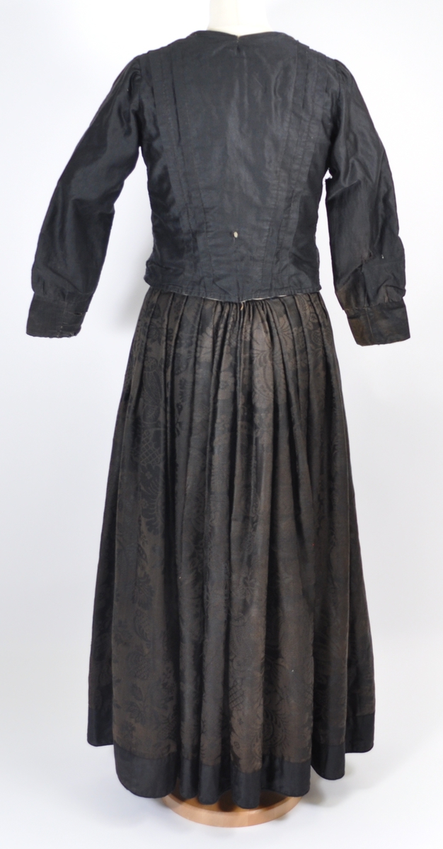 Stakk i brunsvart ulldamask der stoffet i dag er litt falmet, slitt og har noen hull. Stakken er sydd for hånd og maskin, og har blitt bøtt og reparert flere steder. Stakken er skjøtet på og består av sju vevhøyder med jarekant, sydd sammen med attersting med ubleket bomullstråd.  Stakken har et glatt stykke midt på fanghøyden som er 22,5 cm bredt, og foldelagt på begge sider. 28,5 cm lang åpning og en 8 cm bred fold på venstre side av fremstykket. Åpningen har blitt forsterket sekundært med toskafts bomullstøy som er sydd med faldesting med svart sytråd. Stakken har åtte folder foldelagt mot høyre og ti folder foldelagt mot venstre. Midt på bak har stakken et 8 cm bredt felt med rynking. Linningen er av vadmel og trolig sekundær, sydd på med maskin med svart sytråd. Stakken lukkes med to metallhekter og har en metallkrok midt på bak på linningen for å hekte på livet. Stakken har blitt forlenget sekundært med en 8 cm bred kant i svart, glatt bomull, påsydd med maskin med svart sytråd. Dette bomullsstoffet ser ut til å være samme stoff som kjoleliv 6.3.15. Stakken har en 7,2 cm høy skoning i trolig hjemmevevd svart og beige fiskebensmønstret bomull sydd på med maskin og faldesting med svart sytråd. Kantet nede med et 0,5 cm bredt svart kjøpe-ullbånd, sydd på med maskin og faldesting med svart sytråd. Stakken har en sekundær lomme sydd inn i mellom faldene på høyre side. Lommen er 16 cm bred og 21 cm lang, sydd med maskin med svart sytråd, og er i samme stoff som skoningen. Stakken har en metallkrok midt på bak på linningen for å henge på livet, og stakken skal brukes utenpå livet.