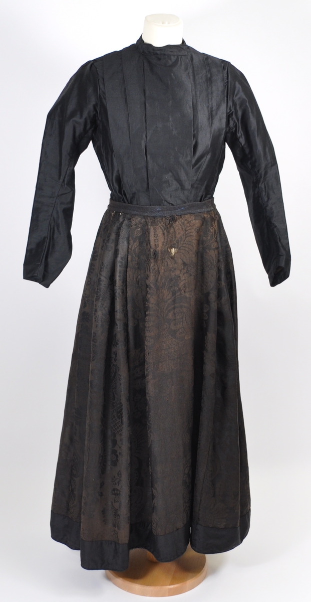 Stakk i brunsvart ulldamask der stoffet i dag er litt falmet, slitt og har noen hull. Stakken er sydd for hånd og maskin, og har blitt bøtt og reparert flere steder. Stakken er skjøtet på og består av sju vevhøyder med jarekant, sydd sammen med attersting med ubleket bomullstråd.  Stakken har et glatt stykke midt på fanghøyden som er 22,5 cm bredt, og foldelagt på begge sider. 28,5 cm lang åpning og en 8 cm bred fold på venstre side av fremstykket. Åpningen har blitt forsterket sekundært med toskafts bomullstøy som er sydd med faldesting med svart sytråd. Stakken har åtte folder foldelagt mot høyre og ti folder foldelagt mot venstre. Midt på bak har stakken et 8 cm bredt felt med rynking. Linningen er av vadmel og trolig sekundær, sydd på med maskin med svart sytråd. Stakken lukkes med to metallhekter og har en metallkrok midt på bak på linningen for å hekte på livet. Stakken har blitt forlenget sekundært med en 8 cm bred kant i svart, glatt bomull, påsydd med maskin med svart sytråd. Dette bomullsstoffet ser ut til å være samme stoff som kjoleliv 6.3.15. Stakken har en 7,2 cm høy skoning i trolig hjemmevevd svart og beige fiskebensmønstret bomull sydd på med maskin og faldesting med svart sytråd. Kantet nede med et 0,5 cm bredt svart kjøpe-ullbånd, sydd på med maskin og faldesting med svart sytråd. Stakken har en sekundær lomme sydd inn i mellom faldene på høyre side. Lommen er 16 cm bred og 21 cm lang, sydd med maskin med svart sytråd, og er i samme stoff som skoningen. Stakken har en metallkrok midt på bak på linningen for å henge på livet, og stakken skal brukes utenpå livet.