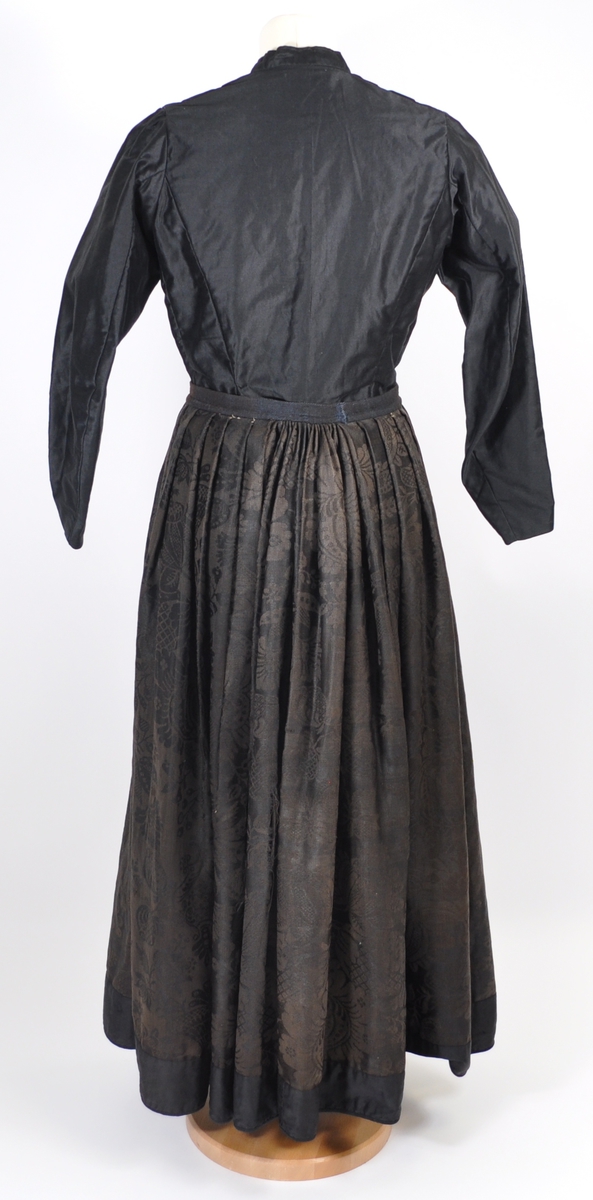 Stakk i brunsvart ulldamask der stoffet i dag er litt falmet, slitt og har noen hull. Stakken er sydd for hånd og maskin, og har blitt bøtt og reparert flere steder. Stakken er skjøtet på og består av sju vevhøyder med jarekant, sydd sammen med attersting med ubleket bomullstråd.  Stakken har et glatt stykke midt på fanghøyden som er 22,5 cm bredt, og foldelagt på begge sider. 28,5 cm lang åpning og en 8 cm bred fold på venstre side av fremstykket. Åpningen har blitt forsterket sekundært med toskafts bomullstøy som er sydd med faldesting med svart sytråd. Stakken har åtte folder foldelagt mot høyre og ti folder foldelagt mot venstre. Midt på bak har stakken et 8 cm bredt felt med rynking. Linningen er av vadmel og trolig sekundær, sydd på med maskin med svart sytråd. Stakken lukkes med to metallhekter og har en metallkrok midt på bak på linningen for å hekte på livet. Stakken har blitt forlenget sekundært med en 8 cm bred kant i svart, glatt bomull, påsydd med maskin med svart sytråd. Dette bomullsstoffet ser ut til å være samme stoff som kjoleliv 6.3.15. Stakken har en 7,2 cm høy skoning i trolig hjemmevevd svart og beige fiskebensmønstret bomull sydd på med maskin og faldesting med svart sytråd. Kantet nede med et 0,5 cm bredt svart kjøpe-ullbånd, sydd på med maskin og faldesting med svart sytråd. Stakken har en sekundær lomme sydd inn i mellom faldene på høyre side. Lommen er 16 cm bred og 21 cm lang, sydd med maskin med svart sytråd, og er i samme stoff som skoningen. Stakken har en metallkrok midt på bak på linningen for å henge på livet, og stakken skal brukes utenpå livet.