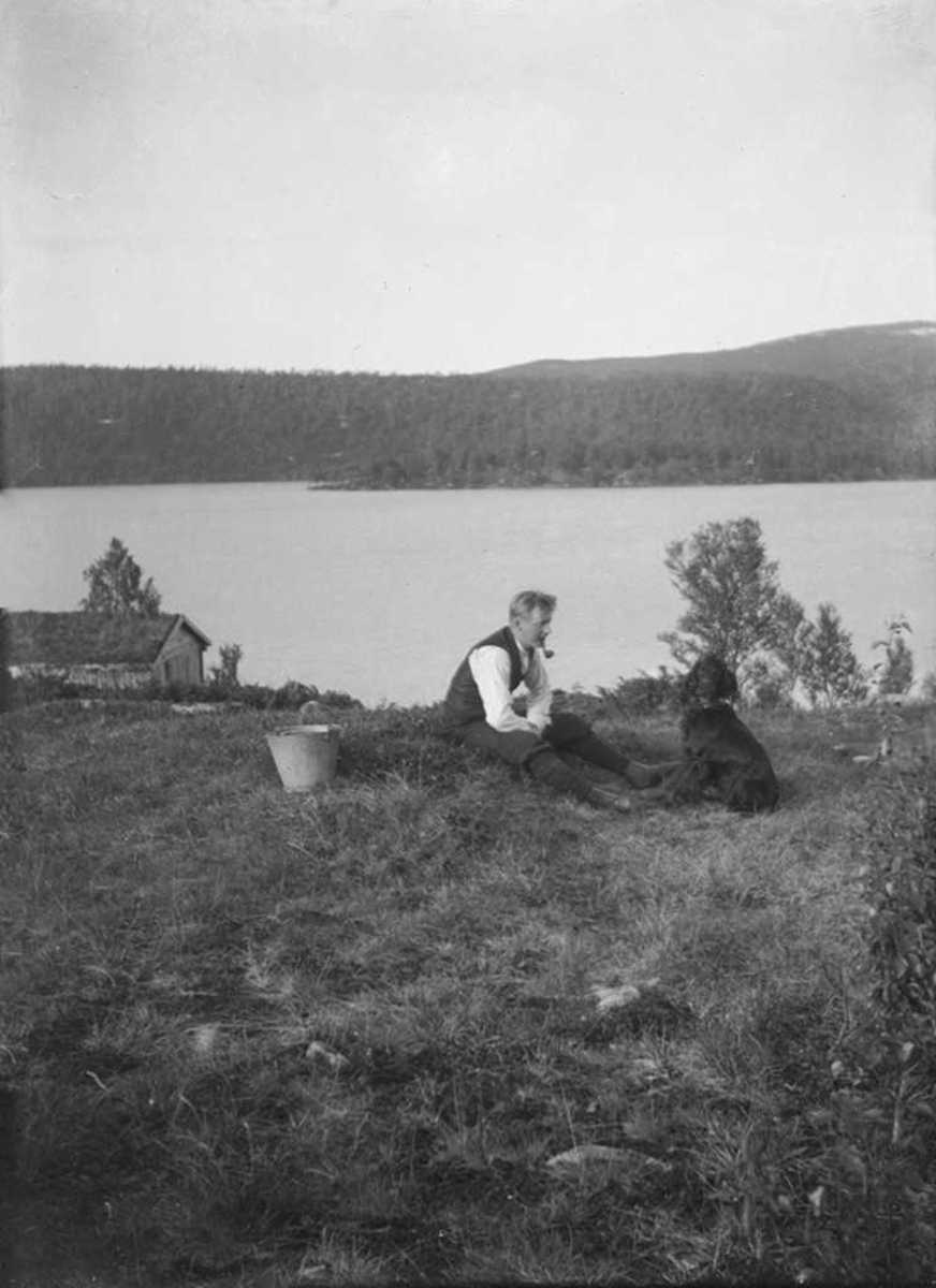 Mann som sitter på gressbakken og røyker pipe. Har en bøtte og en hund med seg. Vann i bakgrunnen.