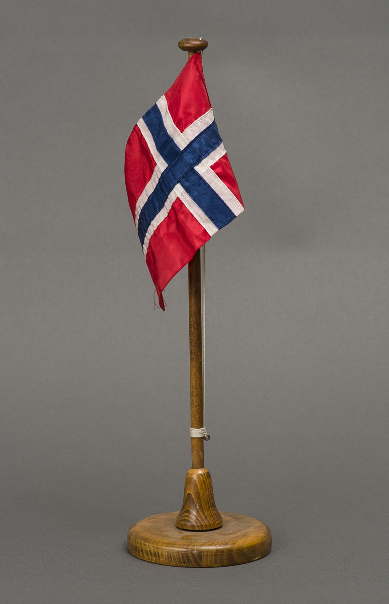 Komplett bordvimpel med stang av tre med rund fot og rektangulær vimpel på snor. Vimpelen, det norske flagg, er rød i bunn med blått kors i midten og hvit kant rundt. Foten er av brunt lakkert tre.