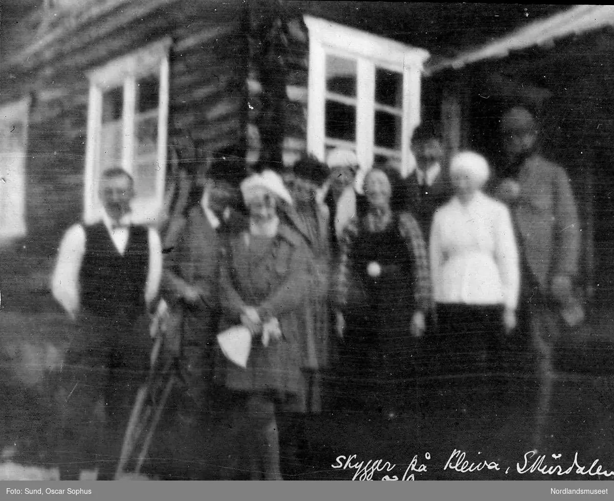 Skursdalen i Hol, Buskerud, Kleiva, 9 personer står foran inngangen til en tømret bygning. Ski står oppstilt ved veggen. Tatt påsken 1919.