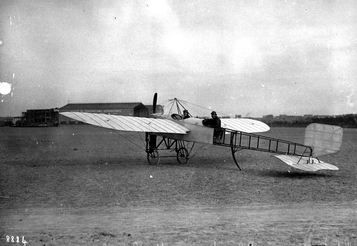 Ett fly på bakken, Bleriot XI.2. To personer, menn, i flyet. Bygninger bak.