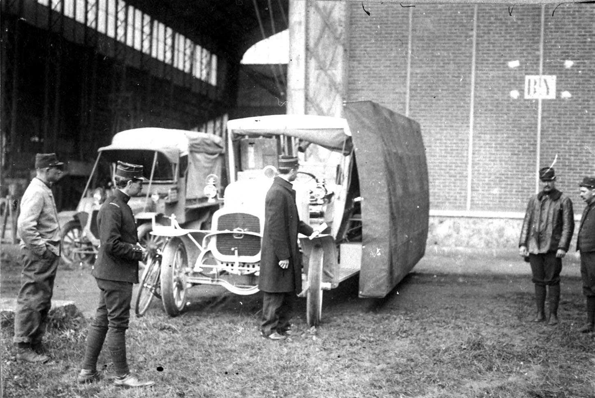 To kjøretøyer foran en hangar. Flyvinge på taket av det ene kjøretøyet. Flere personer, menn i militæruniform ved kjøretøyene.