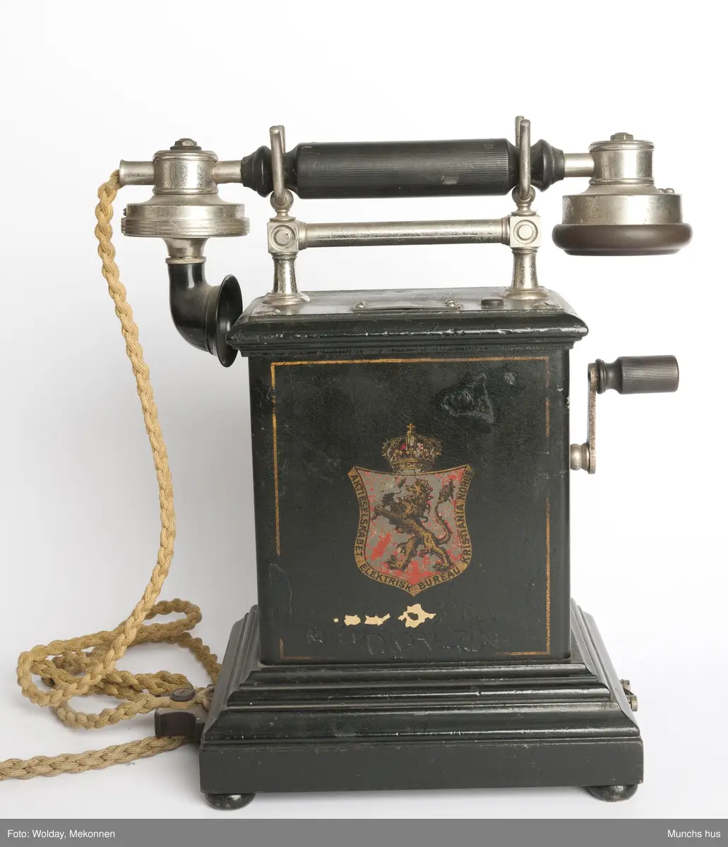 Telefonapparat i sort metall. Foten består av profilerte lister. Sveiv på kassen. På begge sider av kassen er dDen Norske Løve malt i gull og rødt. Rundt riksvåpent er skrevet Aktieselskabet Elektrisk Bureau Kristiania Norge