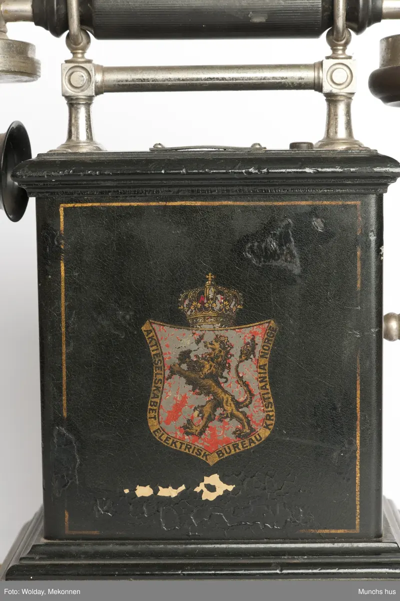 Telefonapparat i sort metall. Foten består av profilerte lister. Sveiv på kassen. På begge sider av kassen er dDen Norske Løve malt i gull og rødt. Rundt riksvåpent er skrevet Aktieselskabet Elektrisk Bureau Kristiania Norge