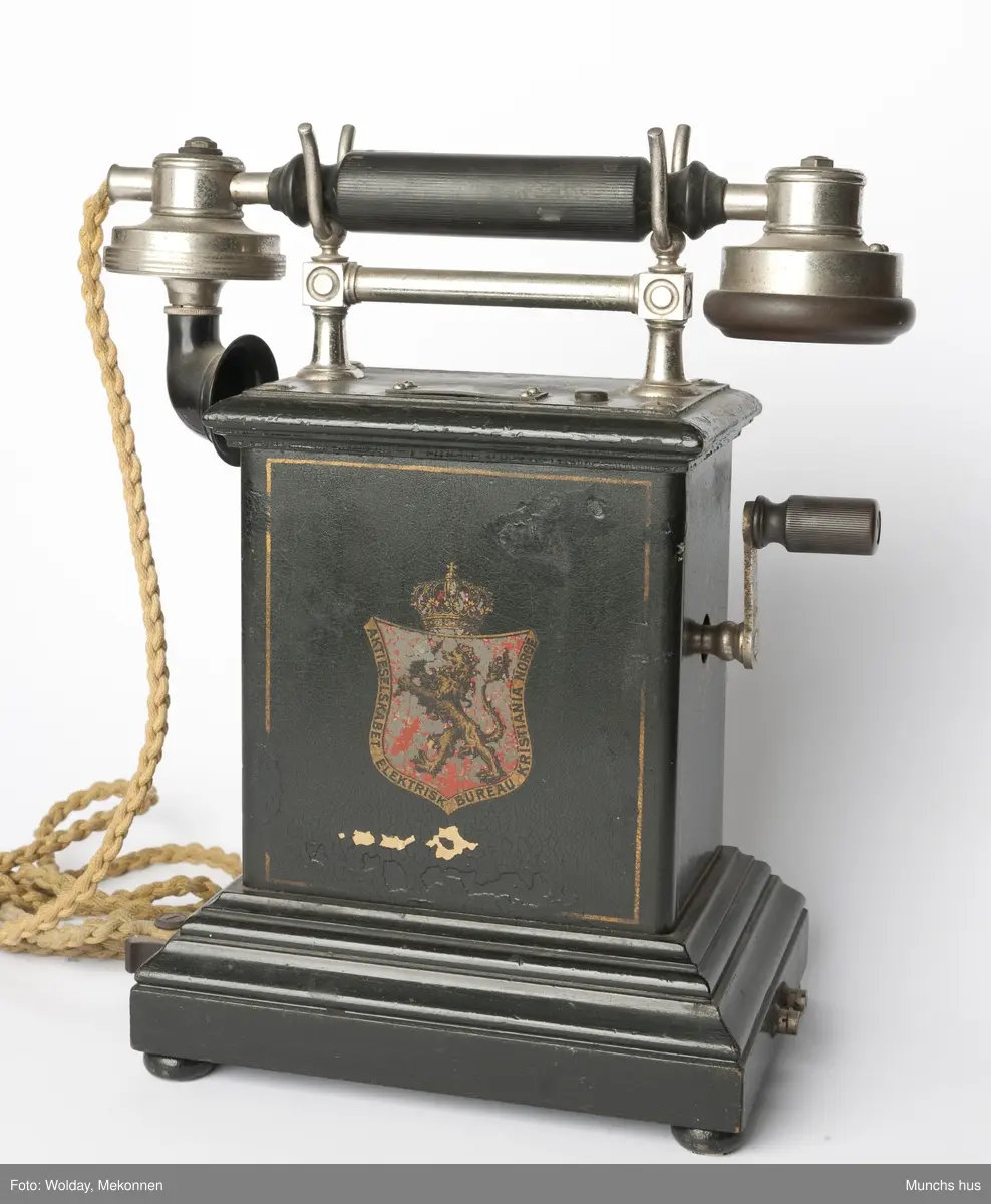 Telefonapparat i sort metall. Foten består av profilerte lister. Sveiv på kassen. På begge sider av kassen er dDen Norske Løve malt i gull og rødt. Rundt riksvåpent er skrevet Aktieselskabet Elektrisk Bureau Kristiania Norge