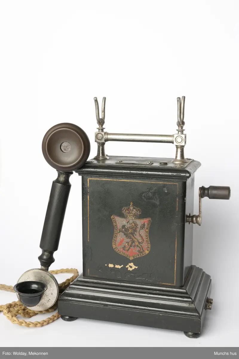 Telefonapparat i sort metall. Foten består av profilerte lister. Sveiv på kassen. På begge sider av kassen er dDen Norske Løve malt i gull og rødt. Rundt riksvåpent er skrevet Aktieselskabet Elektrisk Bureau Kristiania Norge