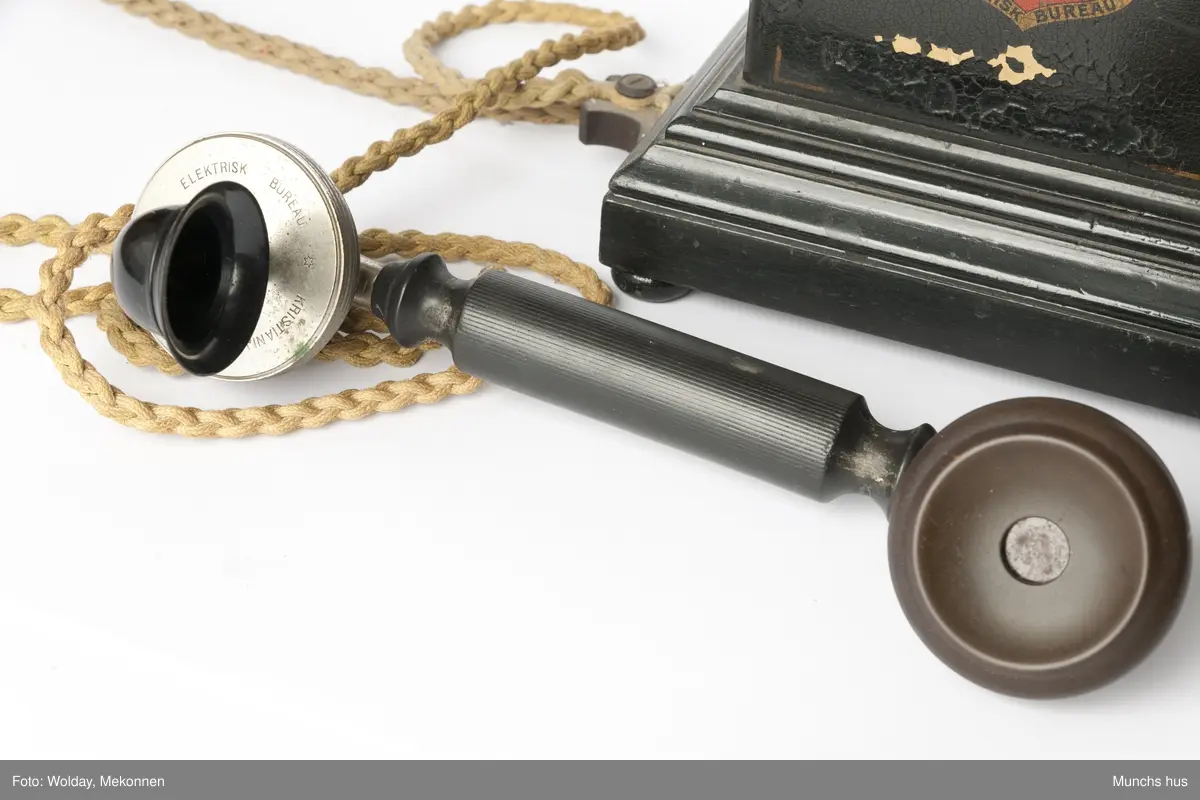 Telefonapparat i sort metall. Foten består av profilerte lister. Sveiv på kassen. På begge sider av kassen er dDen Norske Løve malt i gull og rødt. Rundt riksvåpent er skrevet Aktieselskabet Elektrisk Bureau Kristiania Norge