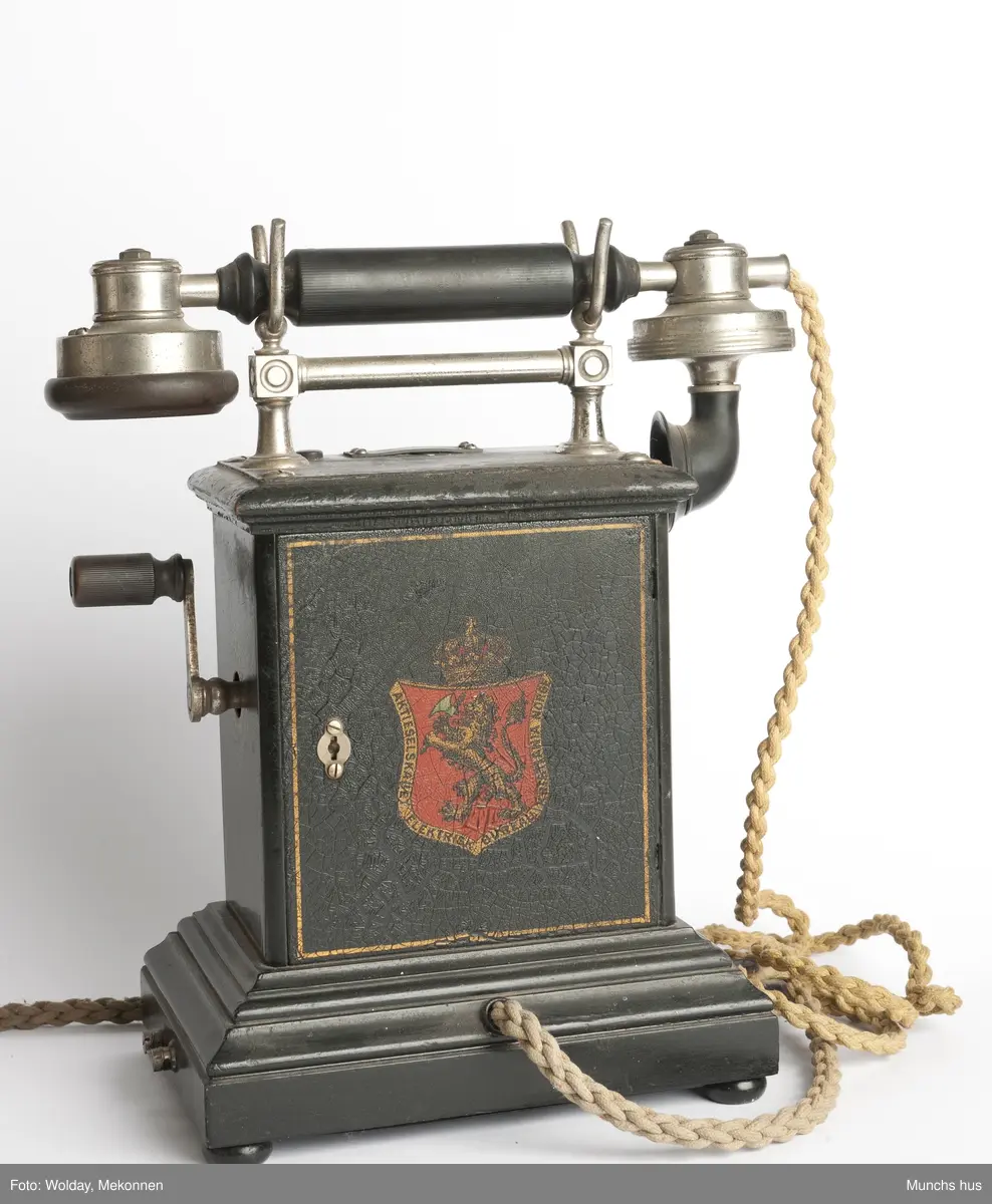 Telefonapparat i sort metall. Foten består av profilerte lister. Sveiv på kassen. På begge sider av kassen er dDen Norske Løve malt i gull og rødt. Rundt riksvåpent er skrevet Aktieselskabet Elektrisk Bureau Kristiania Norge