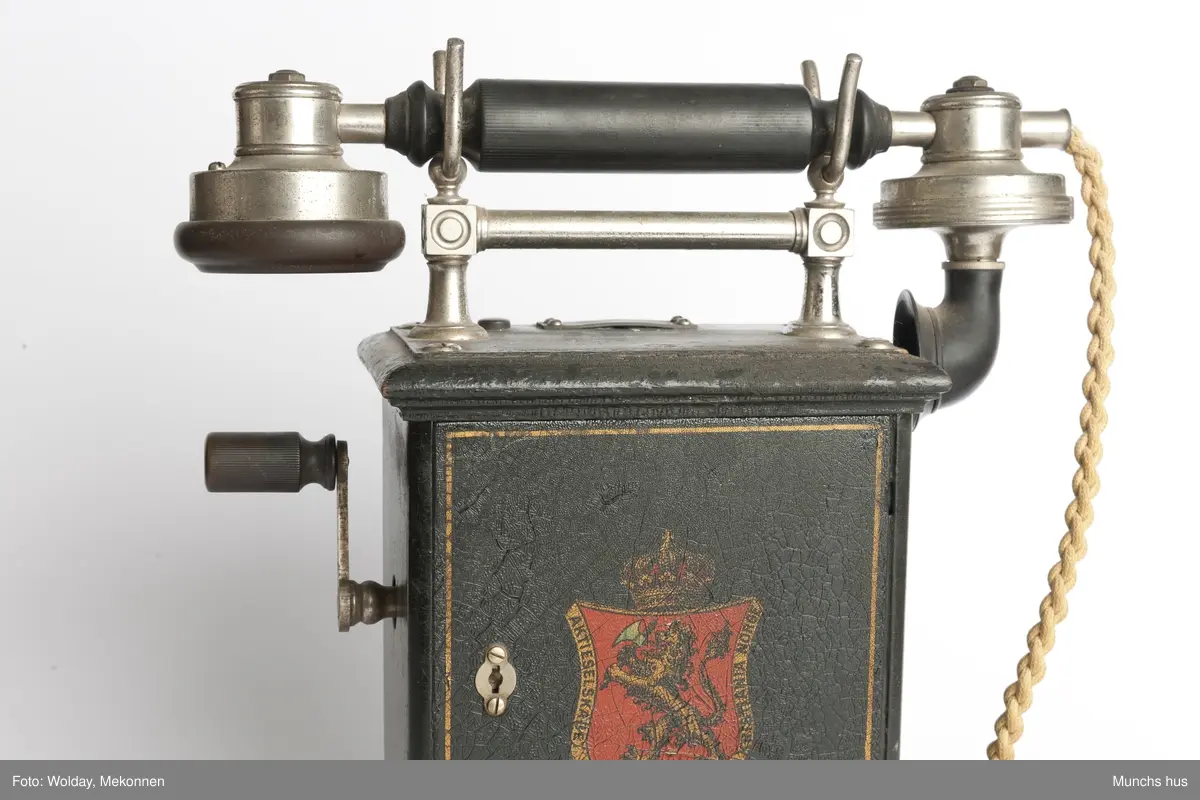 Telefonapparat i sort metall. Foten består av profilerte lister. Sveiv på kassen. På begge sider av kassen er dDen Norske Løve malt i gull og rødt. Rundt riksvåpent er skrevet Aktieselskabet Elektrisk Bureau Kristiania Norge