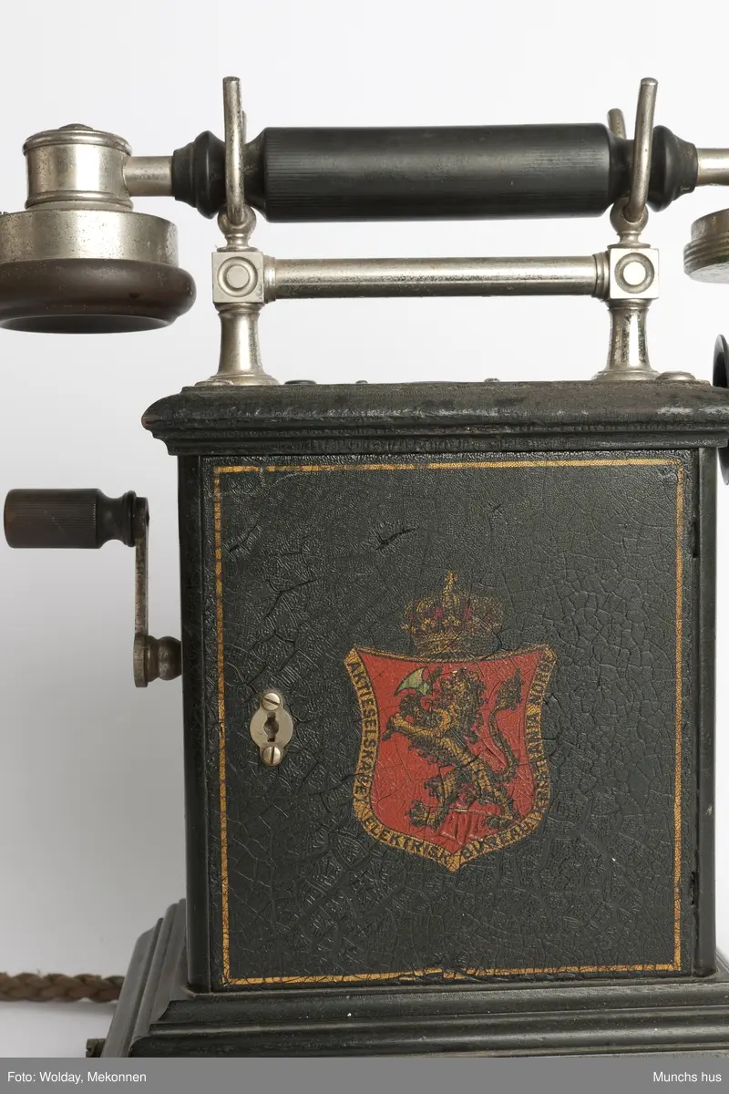 Telefonapparat i sort metall. Foten består av profilerte lister. Sveiv på kassen. På begge sider av kassen er dDen Norske Løve malt i gull og rødt. Rundt riksvåpent er skrevet Aktieselskabet Elektrisk Bureau Kristiania Norge
