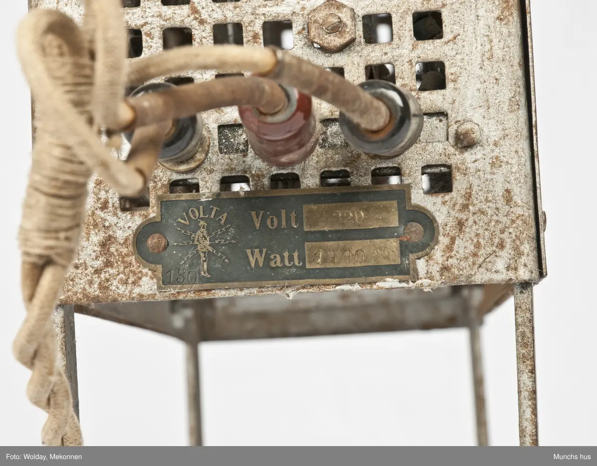 Elektrisk varmeovn. Ovnen yter 1000 Watt - 220 Volt. 
Merket Volta.
Laget av forniklet eller galvanisert jern som er utstyrt med firkantete huller, ca. 1320 stk. den flettede lednikngen er koblet til ovnen med tre plugger, to sorte og en rød. Lednikngen er ca. 290 cm lang og utstyrt med et stort støpsel i porselen. Ovnen har ingen bryter.