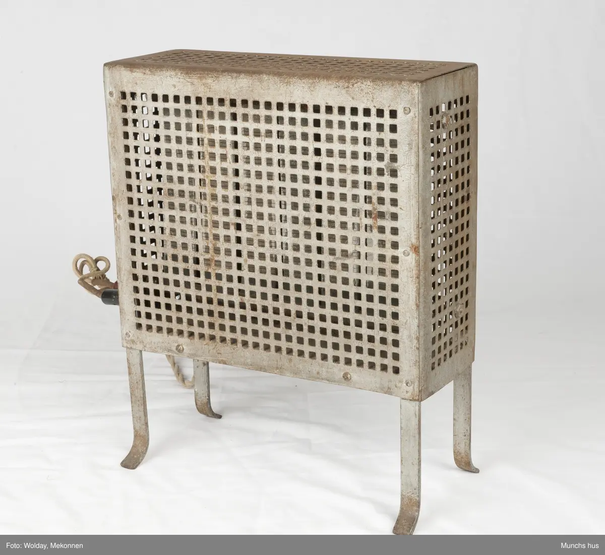 Elektrisk varmeovn. Ovnen yter 1000 Watt - 220 Volt. 
Merket Volta.
Laget av forniklet eller galvanisert jern som er utstyrt med firkantete huller, ca. 1320 stk. den flettede lednikngen er koblet til ovnen med tre plugger, to sorte og en rød. Lednikngen er ca. 290 cm lang og utstyrt med et stort støpsel i porselen. Ovnen har ingen bryter.