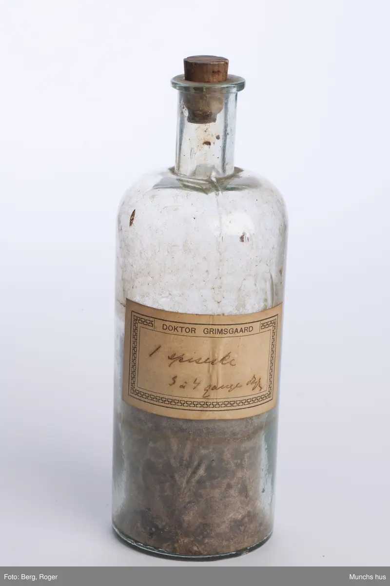 Flaske i klart glass. Medd kork. Noe brunt belegg og grums innvendig.
Målemerker langs flasken med skala 100 - 200 - 300.