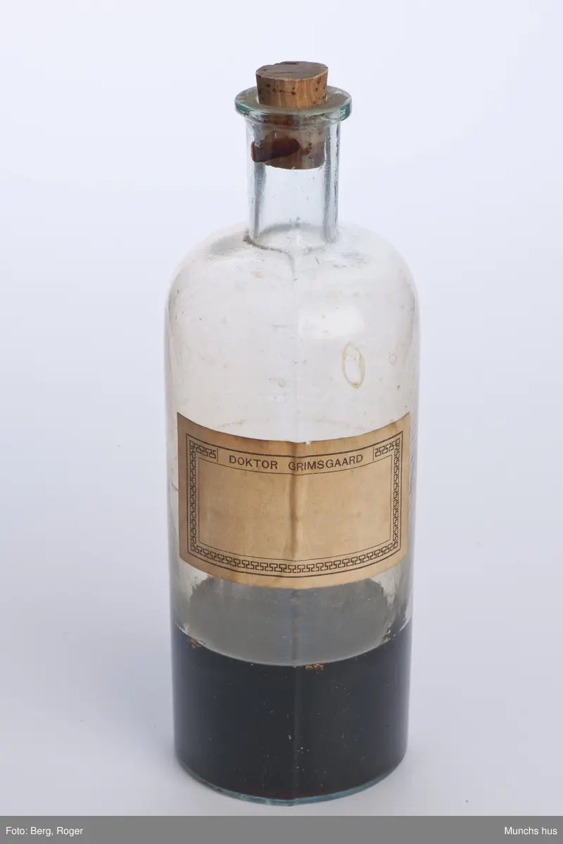 Flaske i klart glass. Med kork. 
Flasken inneholder en rødbrun væske omtrent 1/3 full. Målemerker med skala 100 - 200 - 300 på siden.