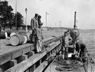 Män fyller på olja (ubåten Hvalen) vid kaj under ubåtsskolan 1911. Alla bär arbetskläder och vegamössa. Runt omkring på kajen syns oljetunnor.